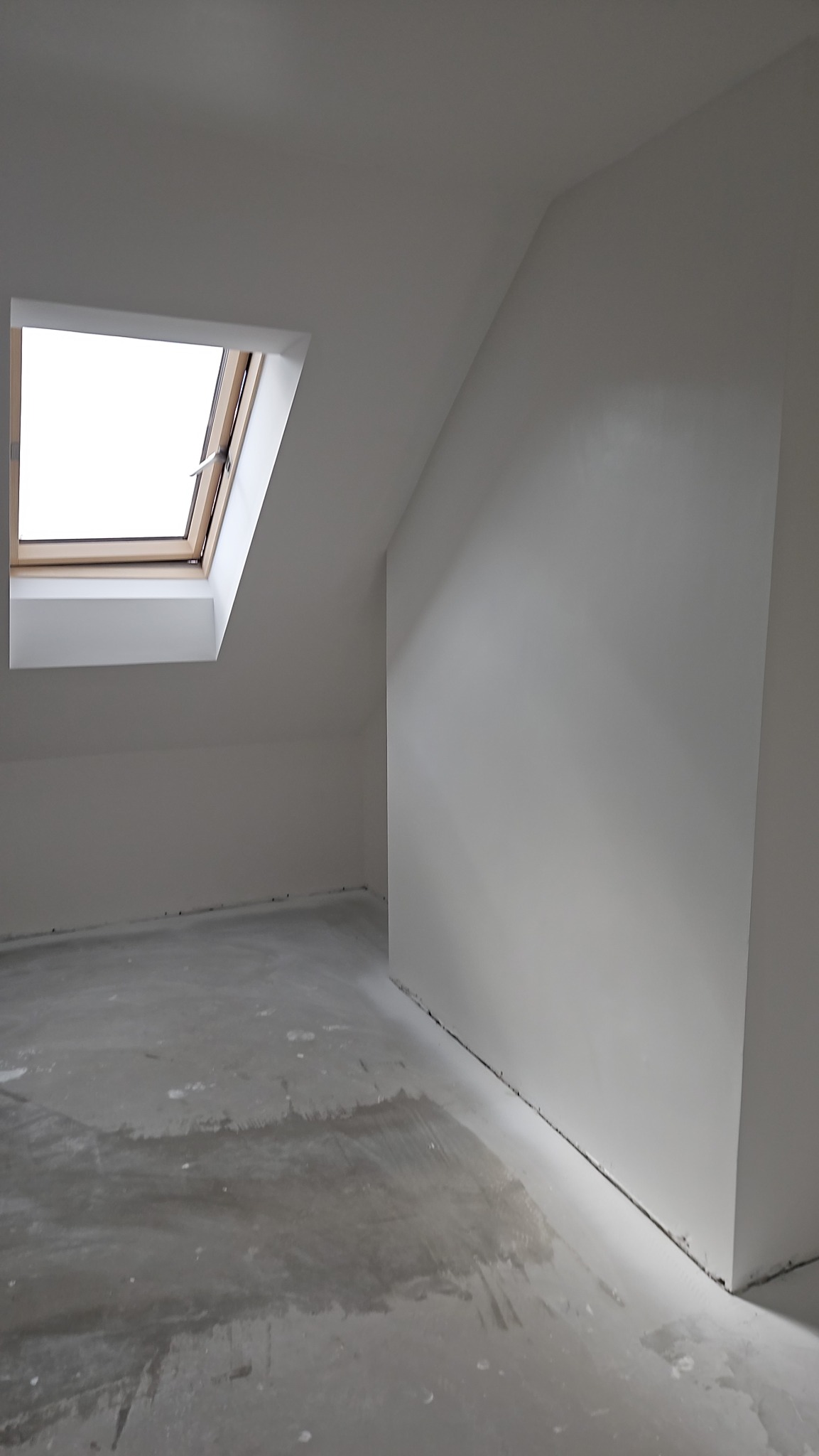 Wnętrze poddasza po szpachlowaniu i malowaniu na biało, widoczne okno dachowe z drewnianą ramą. Podłoga w trakcie prac wykończeniowych.