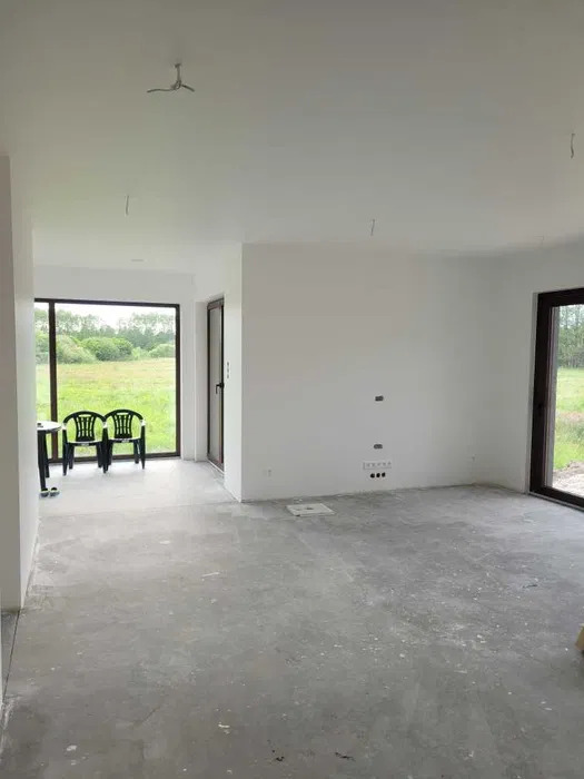 Surowe wnętrze z białymi ścianami i szarą podłogą, widok na zewnątrz przez okno, dwa krzesła przy stole. Przygotowanie do wykończenia, widoczne gniazdka elektryczne.