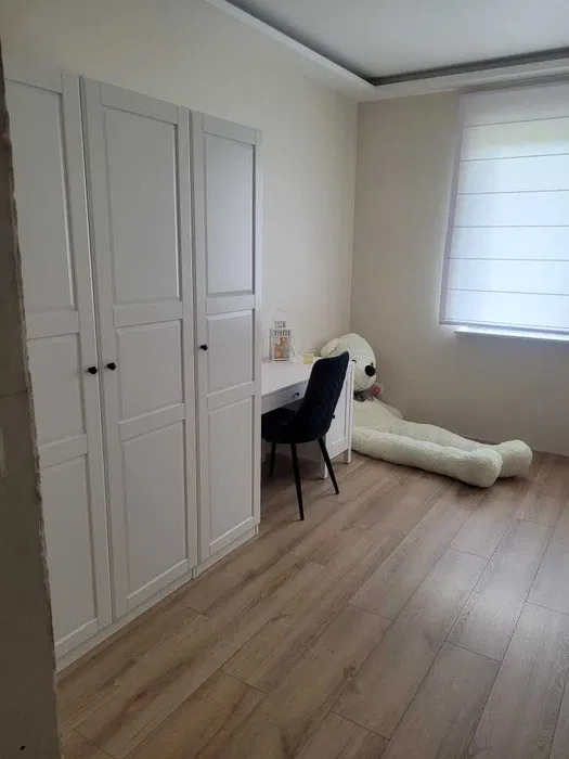 Jasne panele, biała szafa z trzema segmentami i biurko w pokoju. Na podłodze leży duży pluszowy miś. Nowoczesny design.