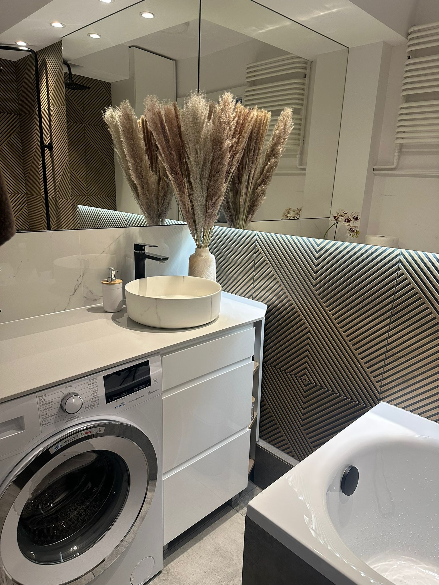 Nowoczesna łazienka z pralką Beko, umywalką nablatową i dekoracyjną ścianą z drewnianych paneli. W lustrze odbija się kabina prysznicowa i grzejnik.