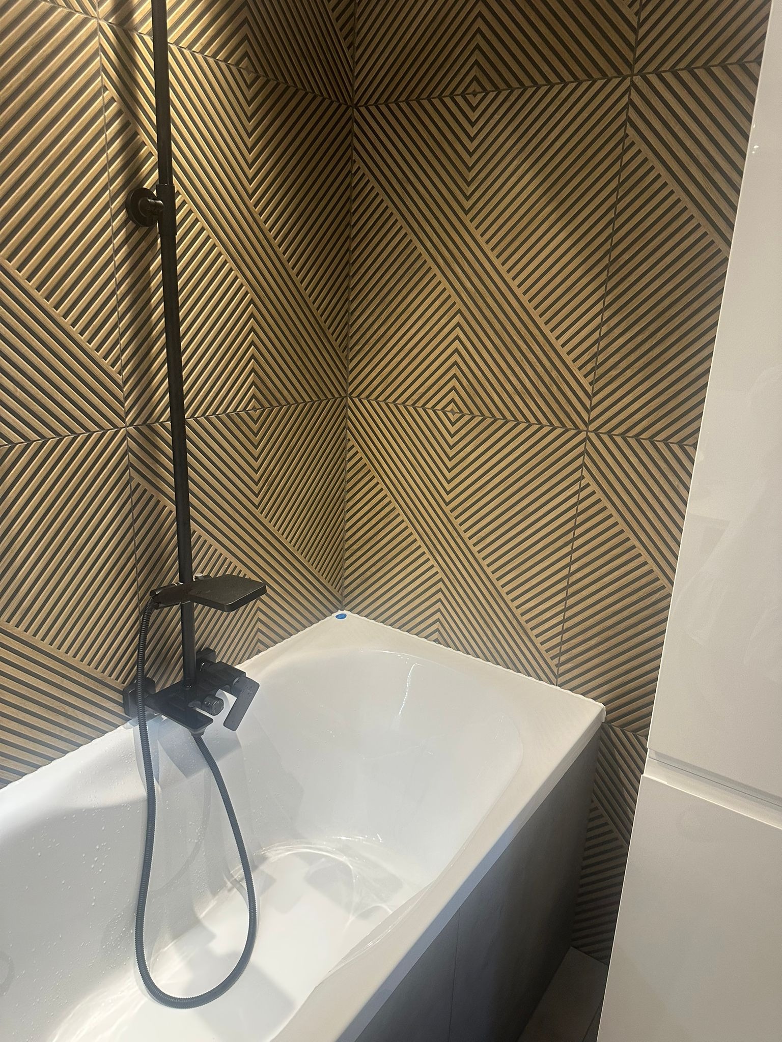 Biała wanna z czarną armaturą na tle ściany z geometrycznym wzorem w odcieniach brązu. Widoczny fragment białej szafki. Nowoczesny design łazienki.