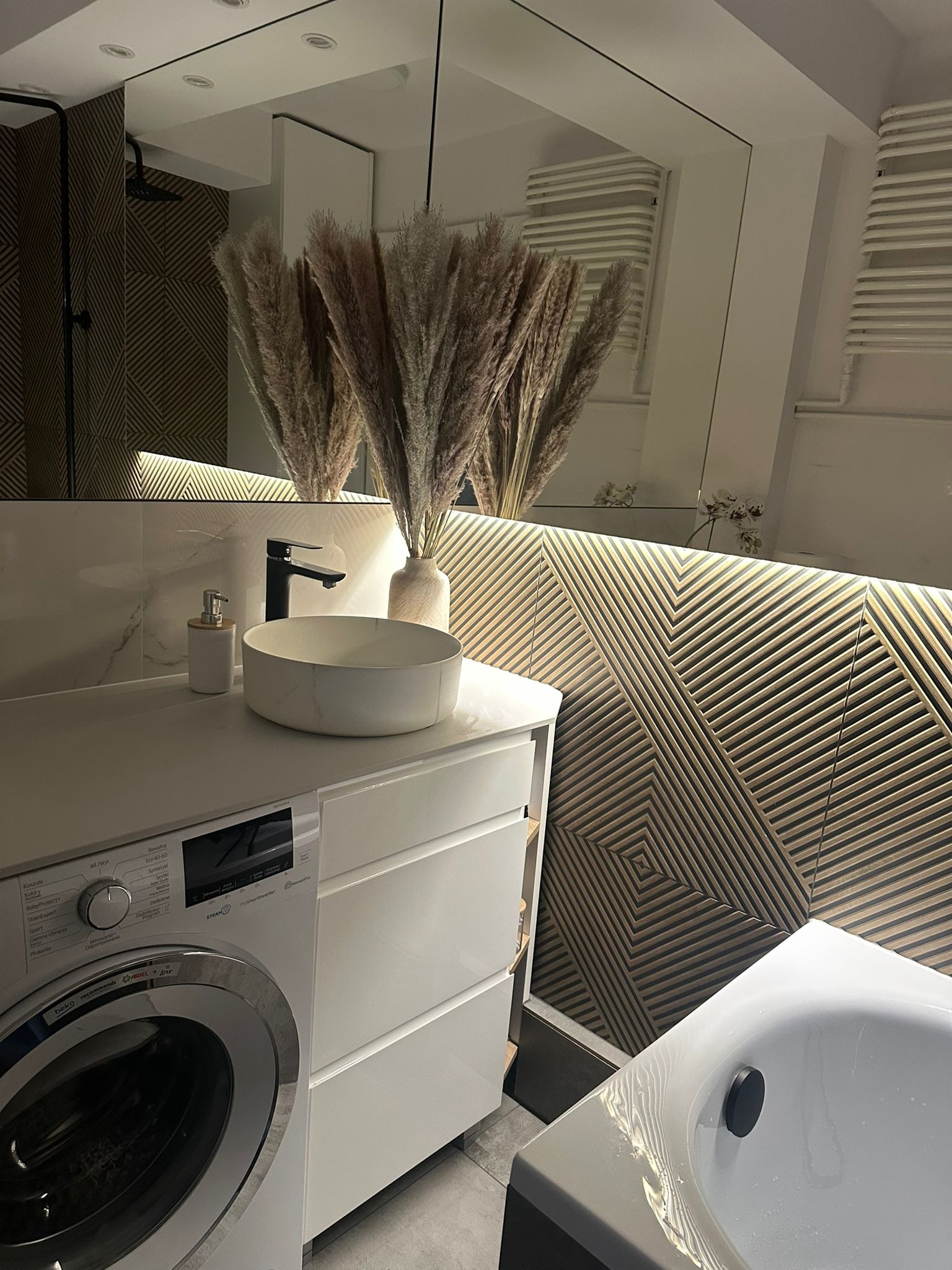 Nowoczesna łazienka z umywalką nablatową, baterią w kolorze czarnym i pralką. Na ścianie dekoracyjne panele 3D z podświetleniem LED. W lustrze odbicie kabiny prysznicowej.