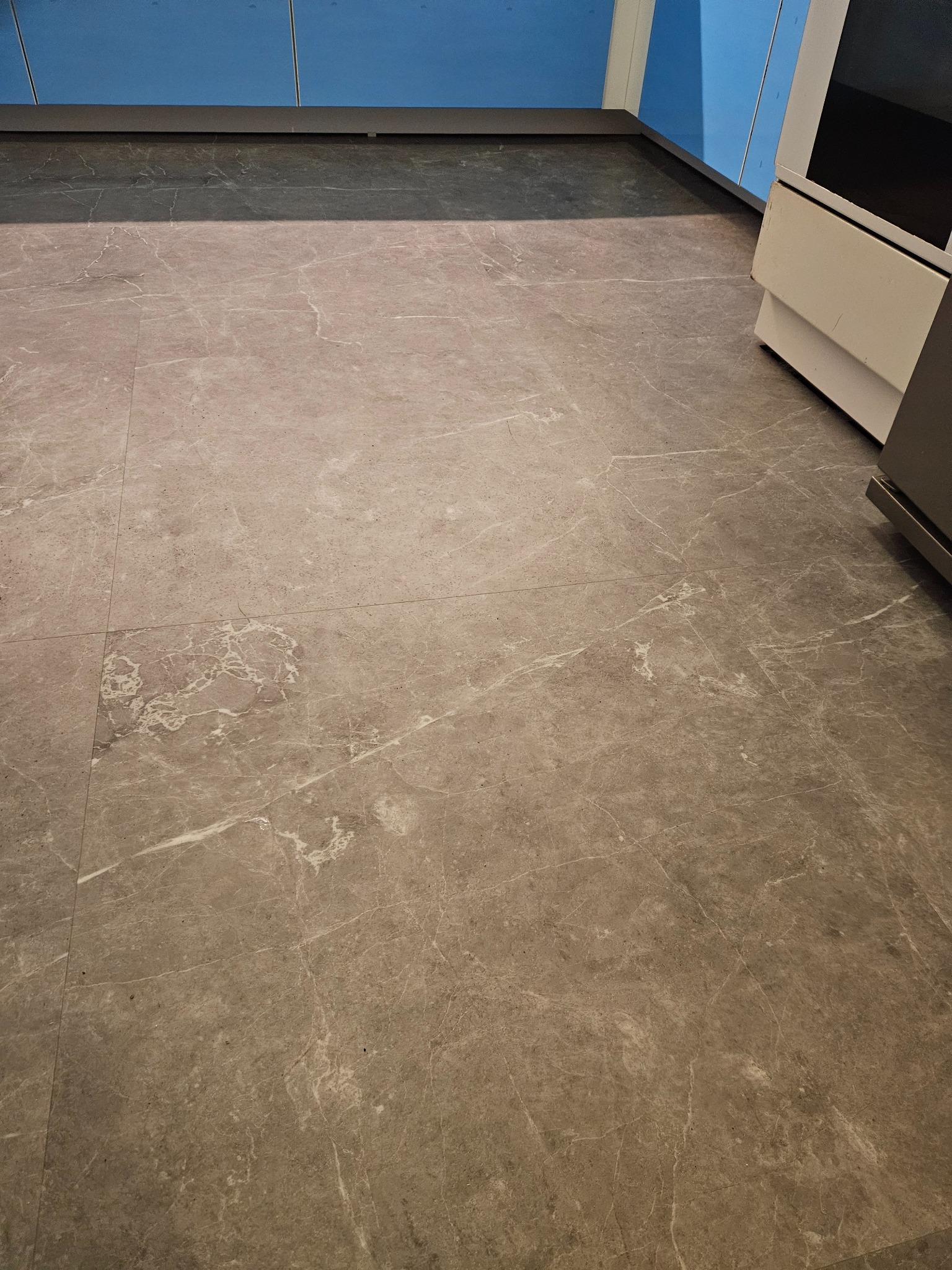 Szara, marmurkowa podłoga w kuchni, z widocznymi płytkami i fragmentem zabudowy kuchennej w tle. Widok z góry, oświetlenie naturalne.