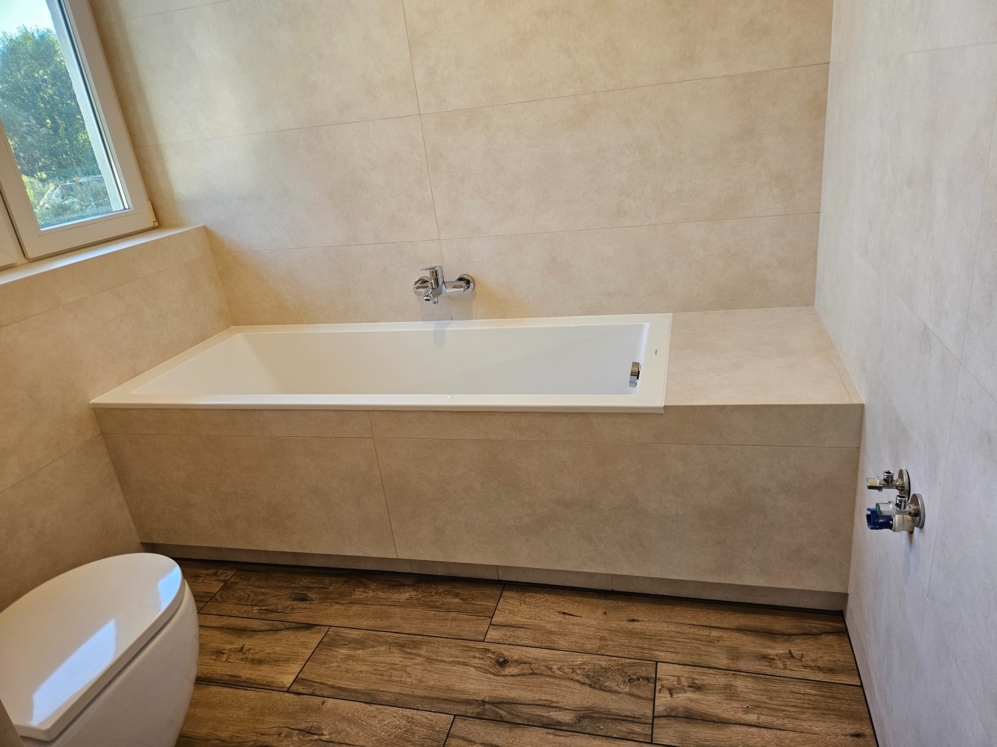 Beżowa łazienka po remoncie: wanna zabudowana płytkami, drewnopodobna podłoga, toaleta. Widoczne zawory na ścianie. Nowoczesny design.