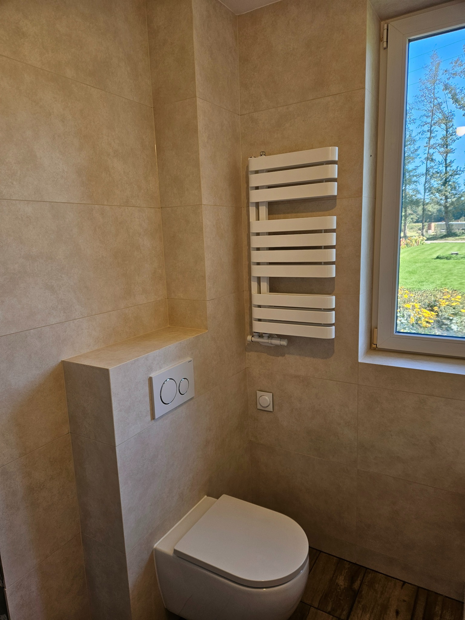Nowoczesna łazienka z beżowymi płytkami, podwieszaną toaletą i grzejnikiem drabinkowym. Widok na zieleń za oknem.