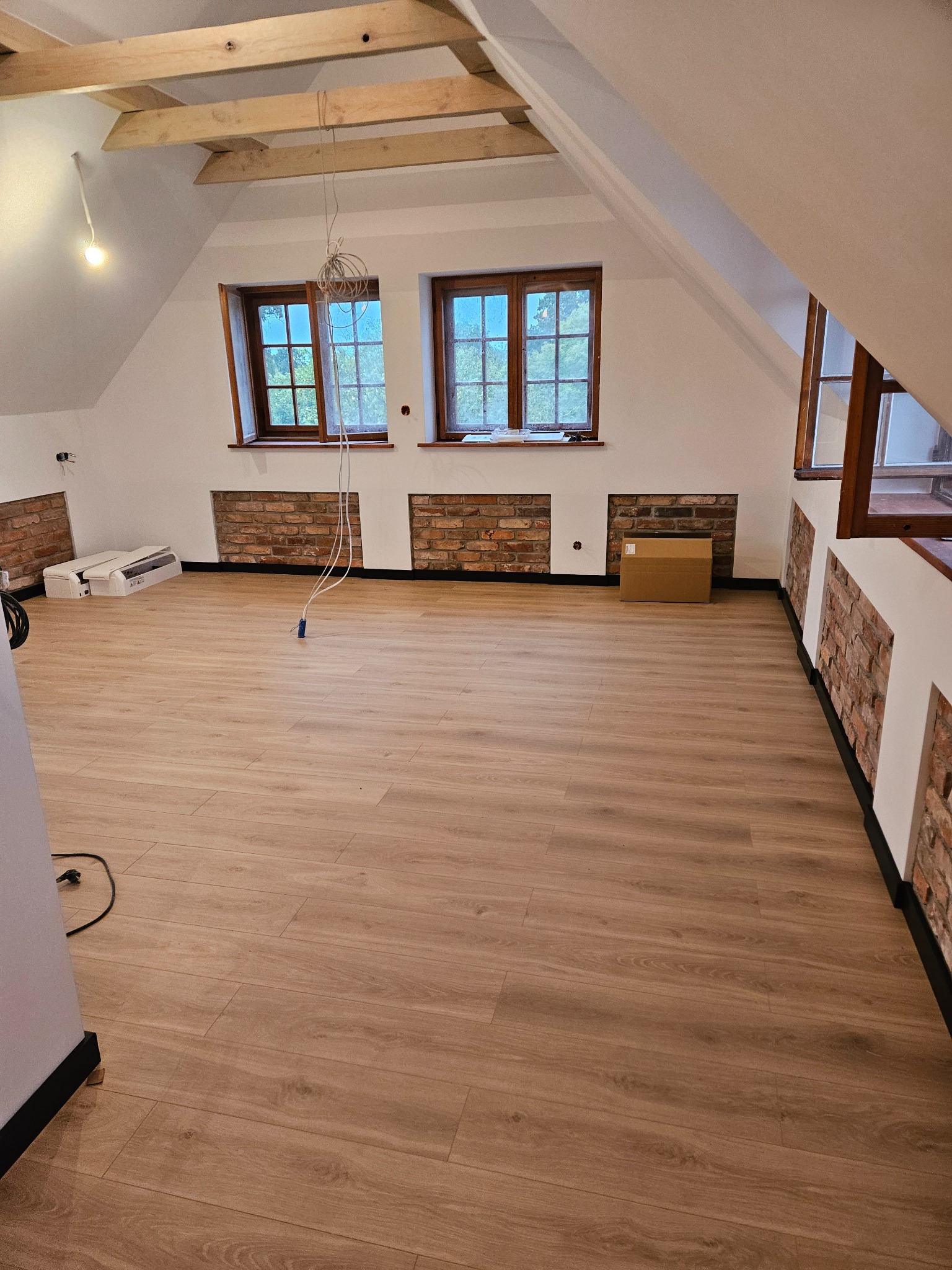 Podłoga z paneli w jasnym odcieniu drewna w nowo wykończonym pokoju na poddaszu z oknami i ceglanymi wnękami. Widoczne drewniane belki stropowe.