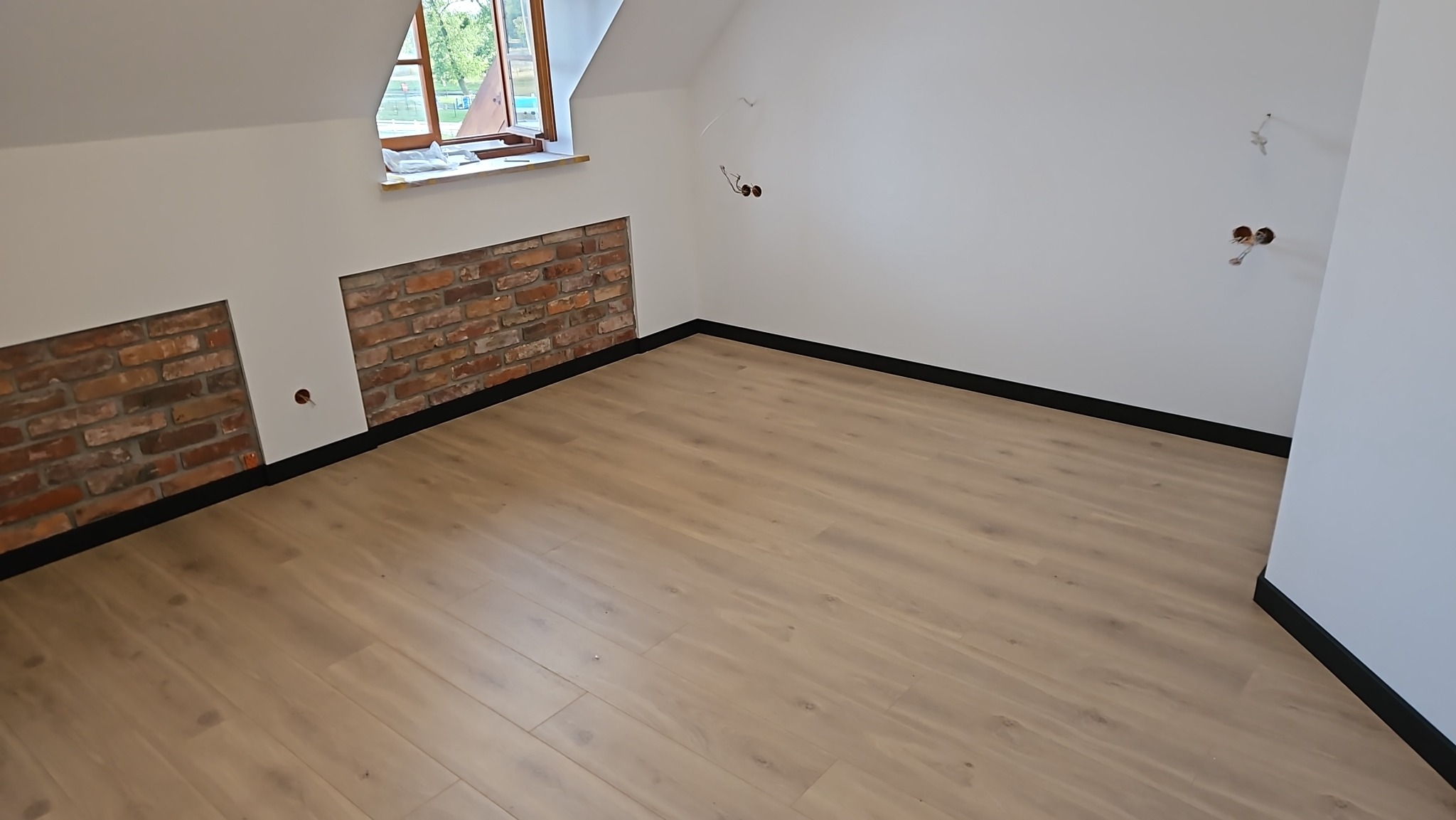 Pokój na poddaszu po remoncie: jasne panele, fragmenty ściany z cegły, białe ściany, okno dachowe. Widoczne gniazdka elektryczne.