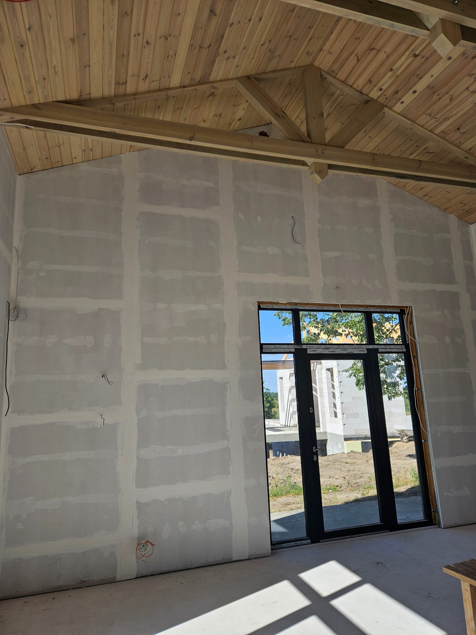 Wnętrze w trakcie prac wykończeniowych: ściany z płyt gipsowo-kartonowych, drewniany sufit z widoczną konstrukcją, duże okno z widokiem na zewnątrz.