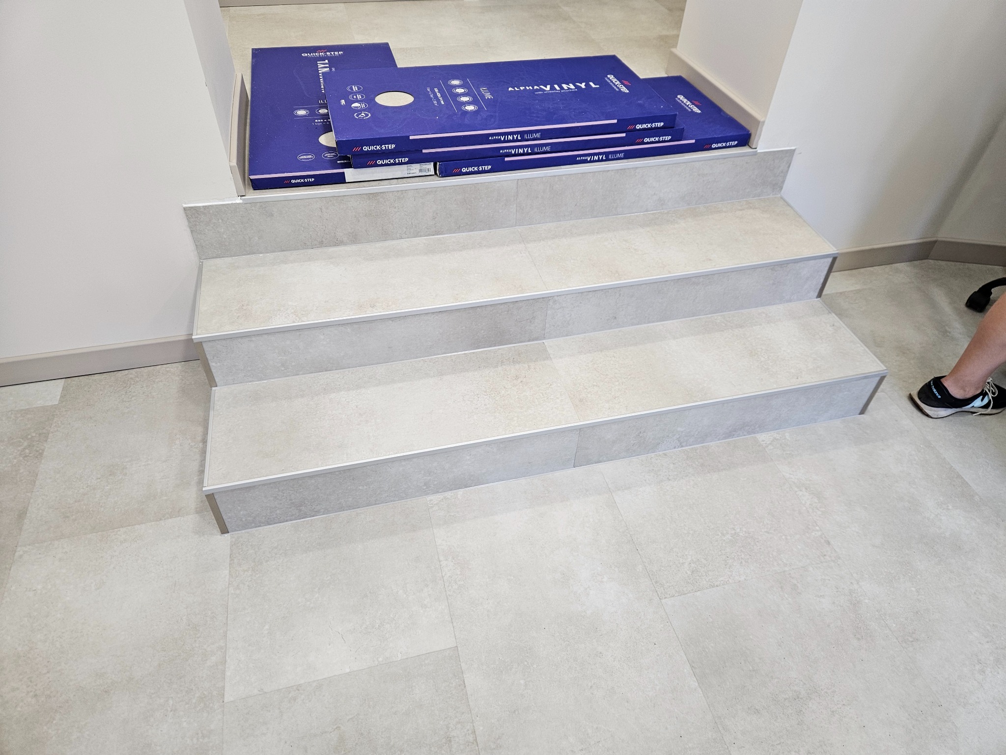 Wykończone schody z płytkami w odcieniach szarości, z widocznymi pudełkami paneli winylowych Quick-Step na górnym stopniu. Minimalistyczne wnętrze, jasne oświetlenie.