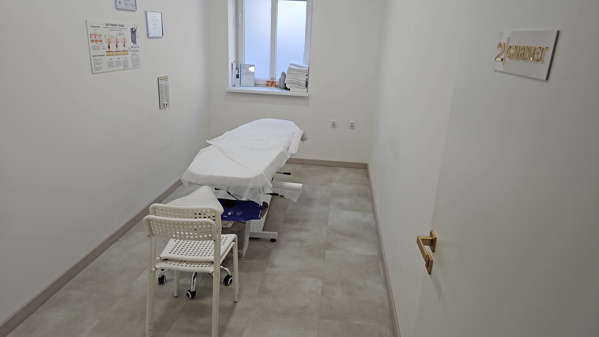 Jasne wnętrze gabinetu kosmetycznego z leżanką przy oknie, białym krzesłem i plakatem informacyjnym na ścianie. Na drzwiach złoty napis 'Gabinet 2'.