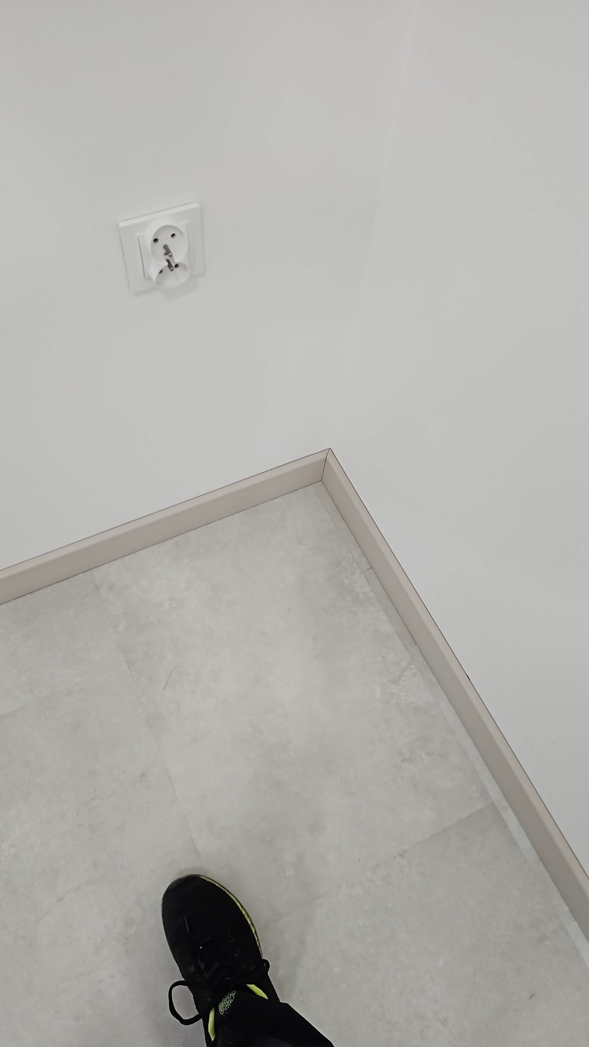 Wykończony narożnik podłogi z listwami przypodłogowymi, widoczny fragment buta osoby fotografującej i gniazdko elektryczne na ścianie.