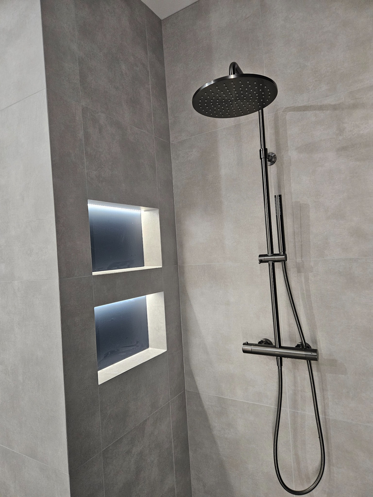 Nowoczesna kabina prysznicowa z deszczownicą i wnękami podświetlonymi LED, wykończona szarą płytką imitującą beton. Minimalistyczny design, eleganckie detale.