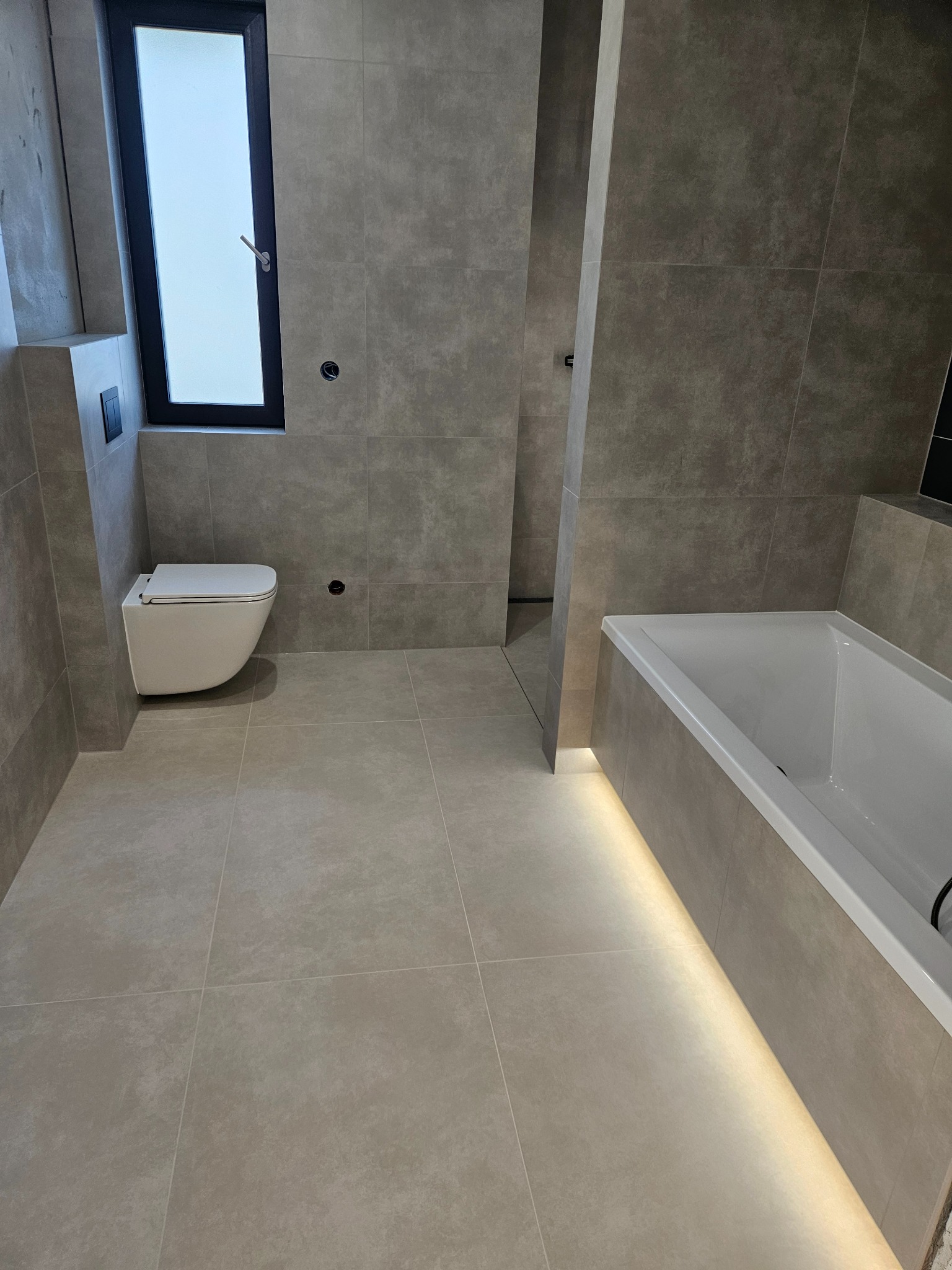 Wykończona łazienka w szarościach z podświetlaną wanną, wiszącą toaletą i wąskim oknem. Minimalistyczny design z dużymi płytkami i subtelnym oświetleniem.