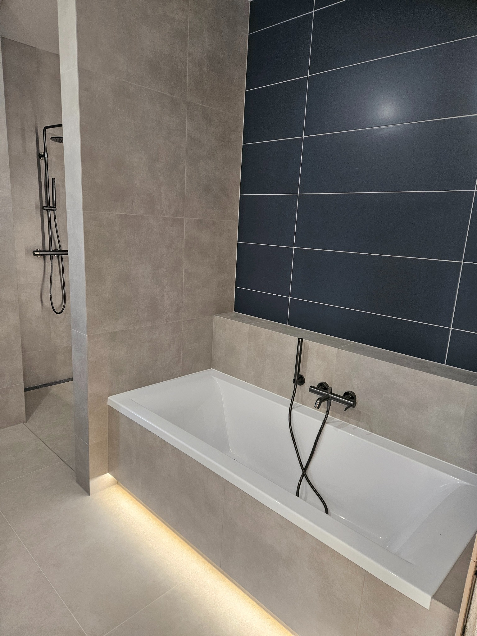 Nowoczesna łazienka z wanną, czarną armaturą i podświetleniem LED. Szare płytki i ciemna ściana z płytek tworzą elegancki kontrast. Widoczny prysznic typu walk-in.