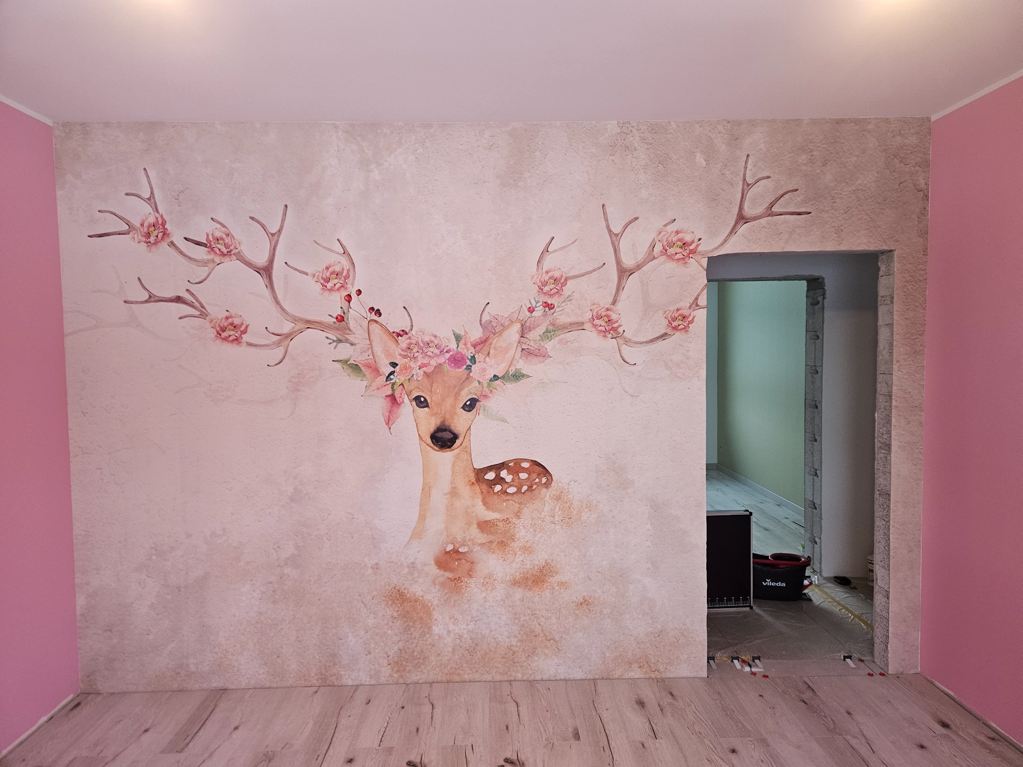 Ściana z dekoracyjną, ręcznie malowaną grafiką jelenia z kwiatami na porożu, w pokoju z różowymi ścianami i drewnianą podłogą. Widoczne narzędzia malarskie.