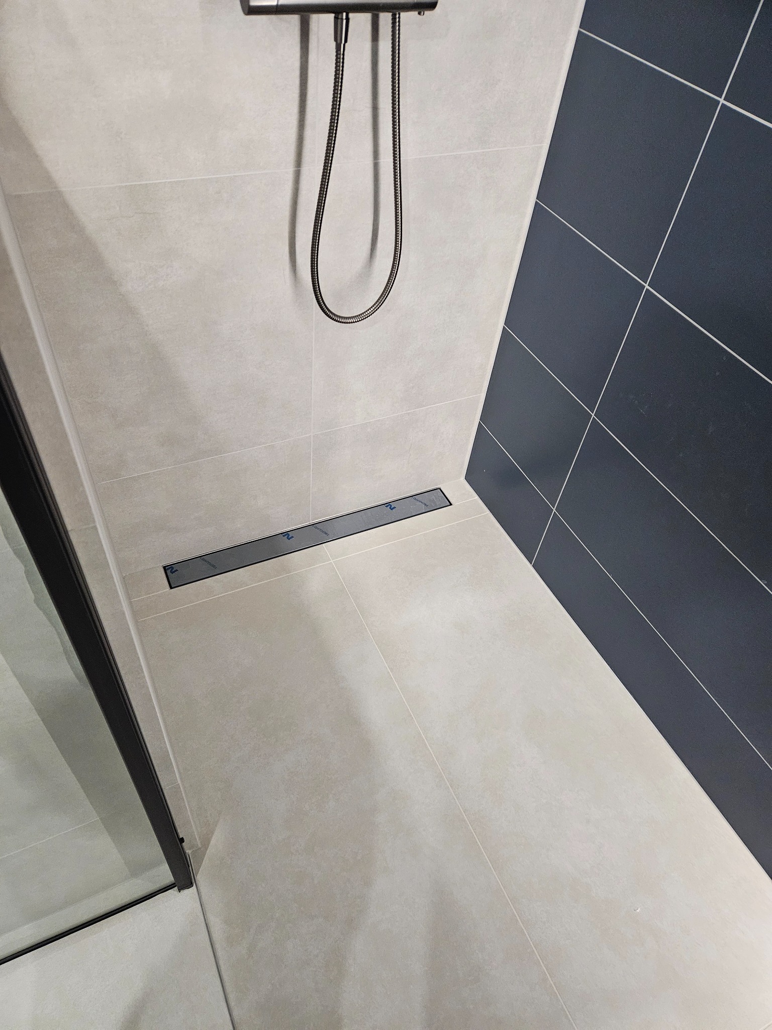 Nowoczesna kabina prysznicowa z odpływem liniowym i grafitową ścianą z płytek. Minimalistyczny design, jasne płytki podłogowe i chromowana armatura.