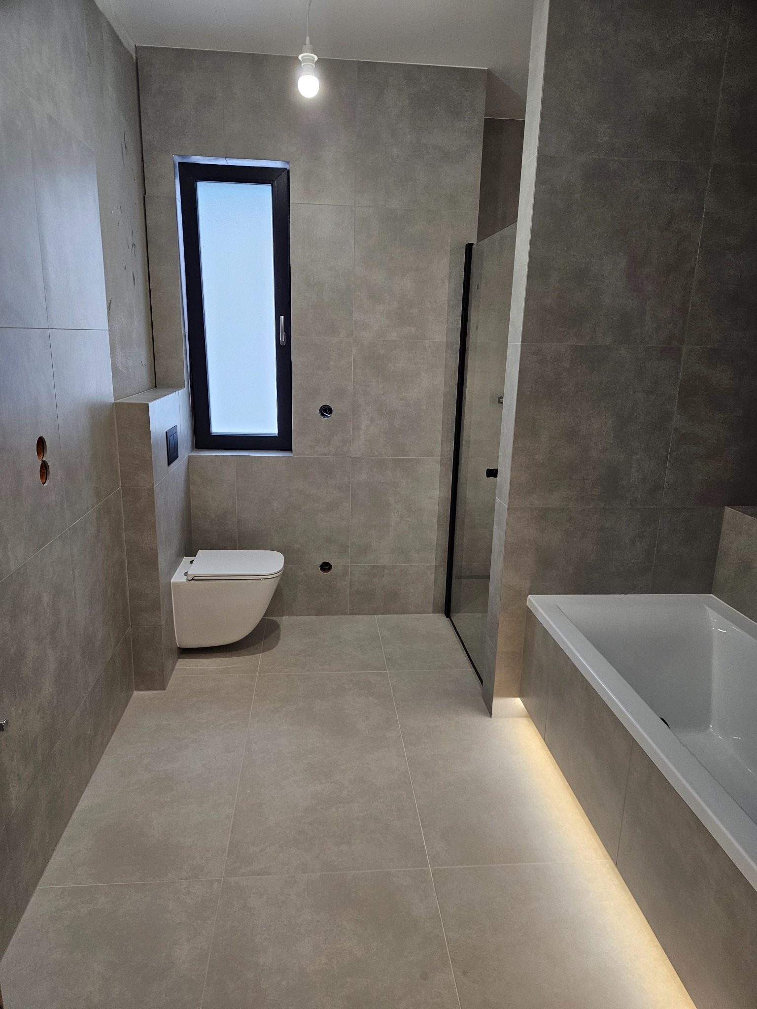 Nowoczesna łazienka w szarościach z podświetlaną wanną i wiszącą toaletą. Minimalistyczny design, czarne ramy okna i kabiny prysznicowej. Ukończone wykończenie.