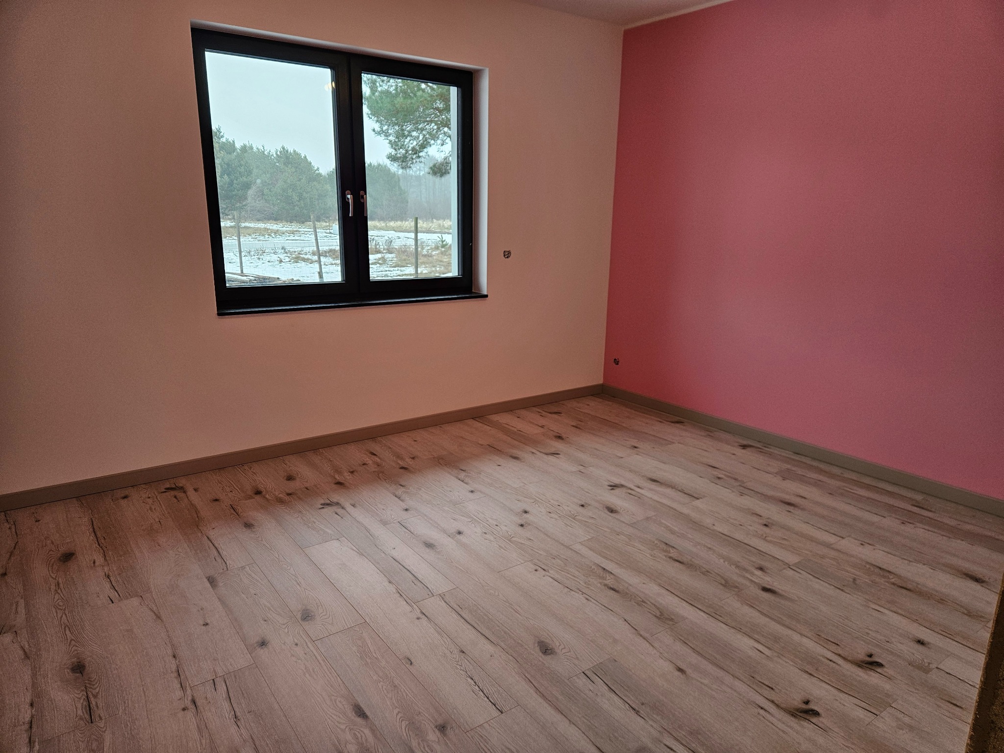 Pokój po remoncie: drewniane panele podłogowe, ściana pomalowana na różowo, okno z widokiem na zimowy krajobraz. Widoczne listwy przypodłogowe.