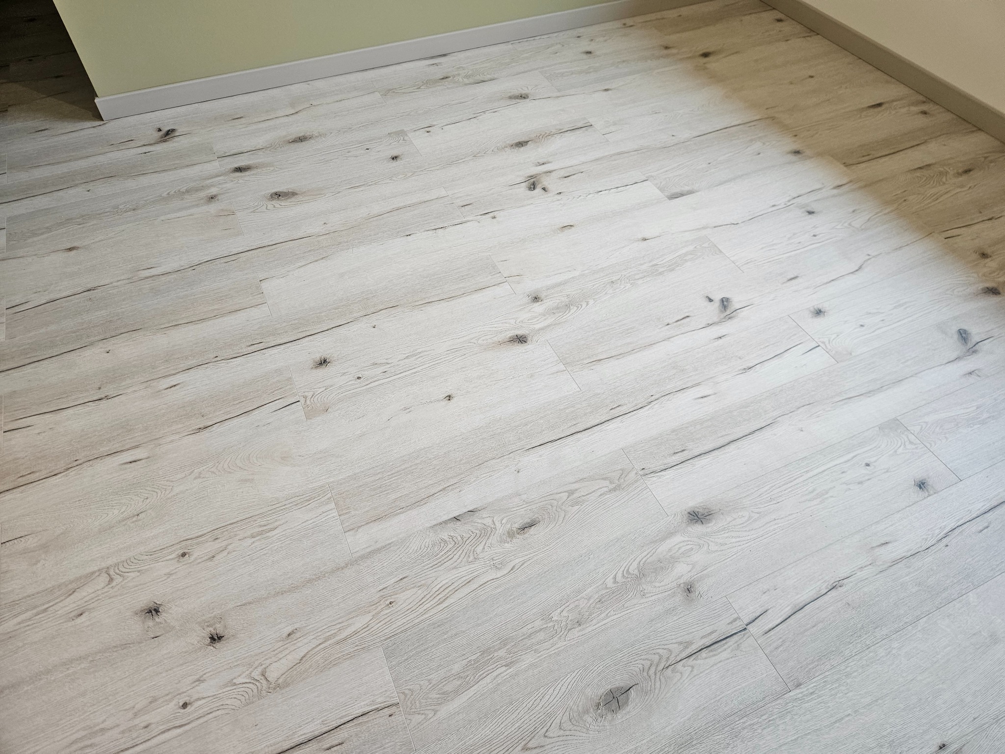 Jasne panele podłogowe z wyraźnym wzorem drewna, ułożone wzdłuż ściany w pokoju z zielonkawą ścianą i białą listwą przypodłogową. Widok z góry, ukośny kadr.