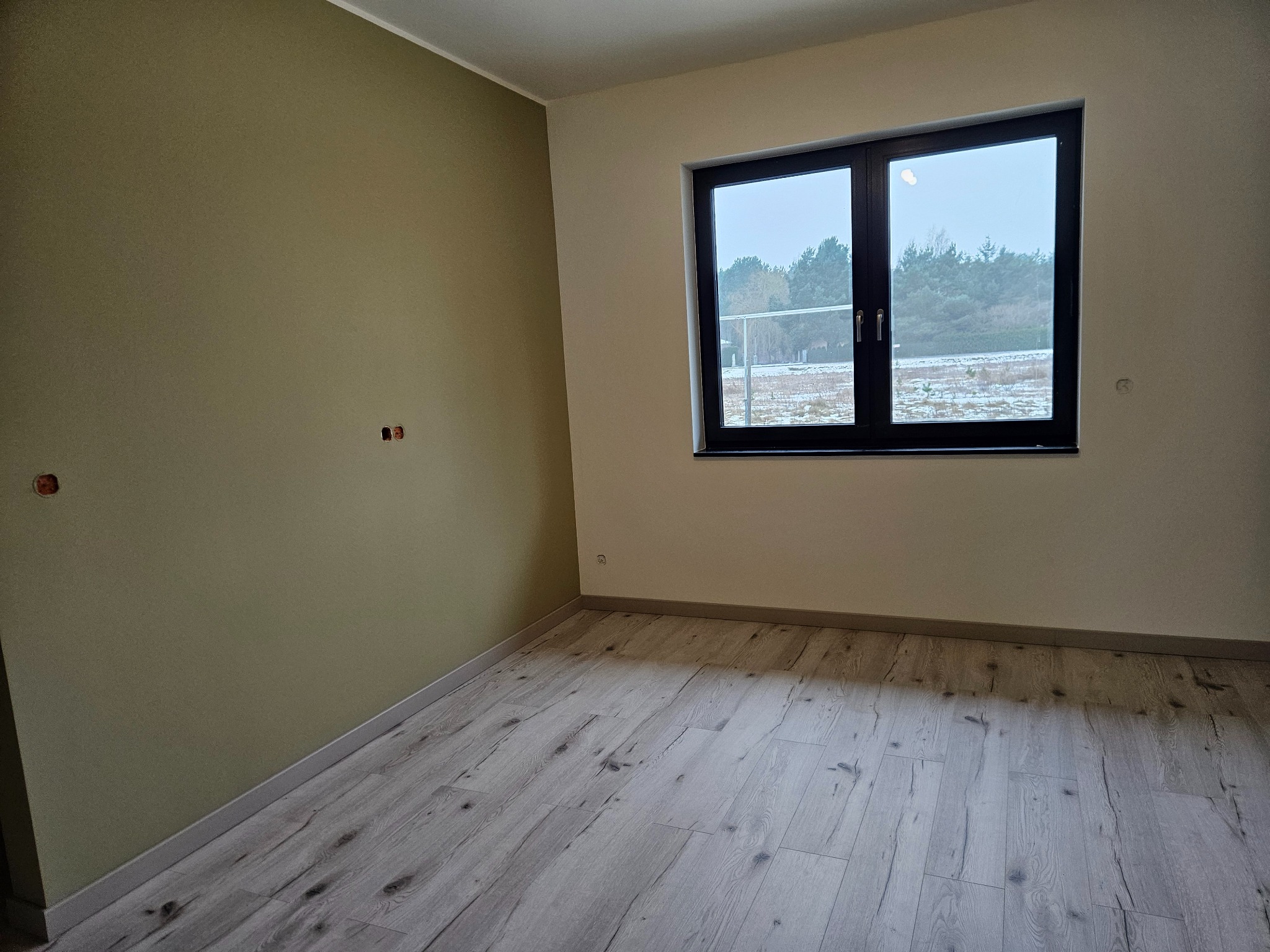 Wnętrze pokoju po malowaniu ścian i ułożeniu paneli podłogowych w Zielonce. Widoczne gładkie ściany w odcieniach beżu i zieleni oraz jasne panele imitujące drewno.