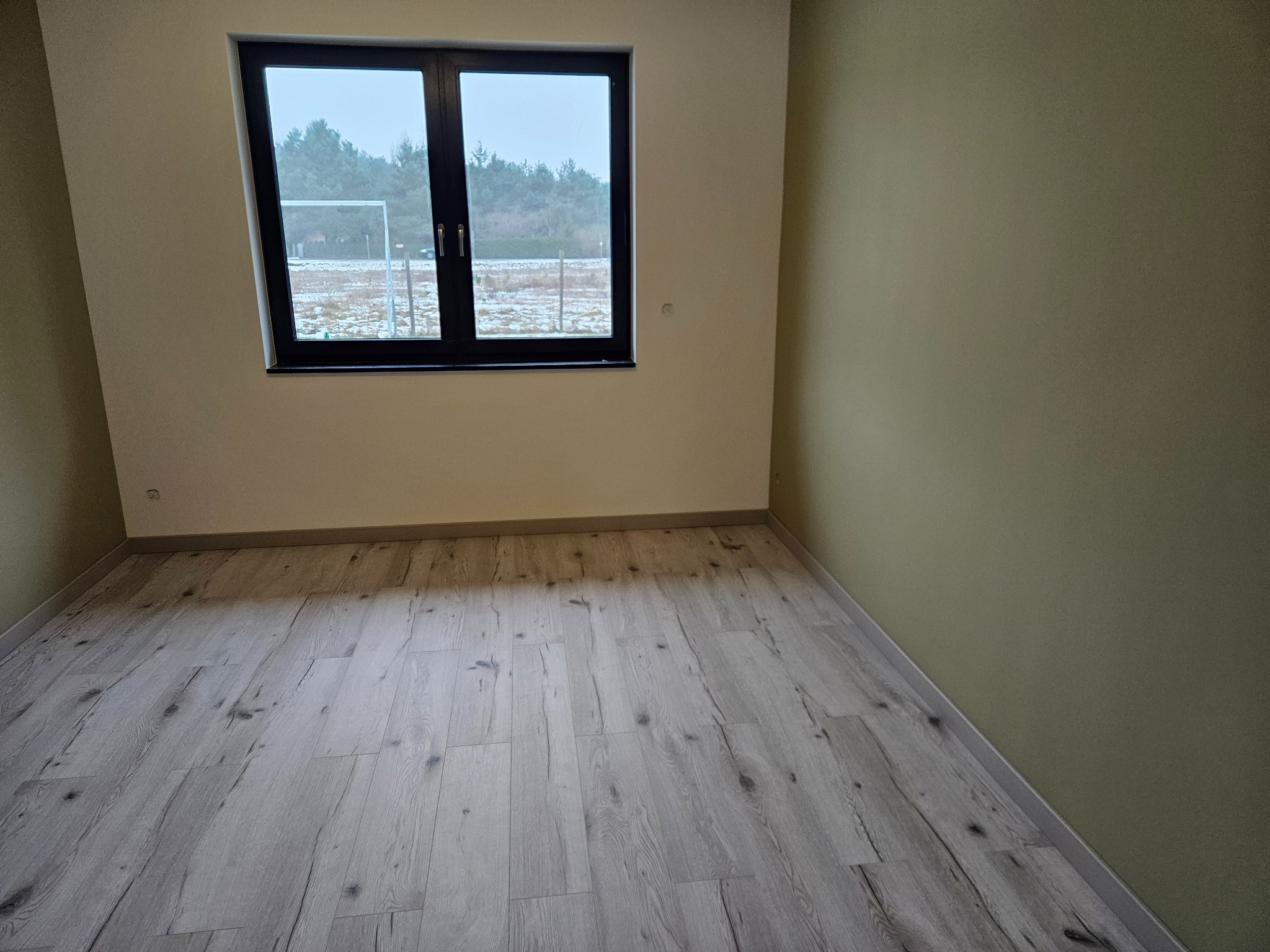 Jasne panele podłogowe w pokoju z oknem na zewnątrz, ściany w odcieniach beżu i zieleni. Widok na boisko sportowe za oknem. Minimalistyczne wykończenie wnętrza.