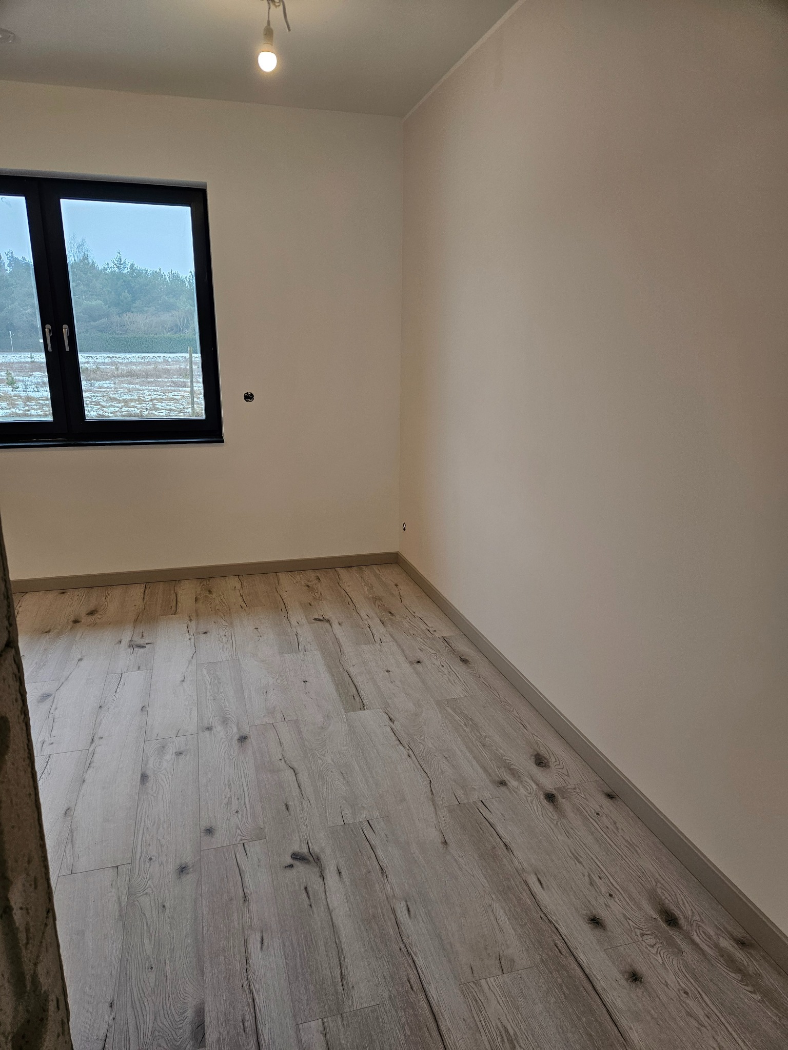 Jasne panele podłogowe w pokoju z oknem i świeżo pomalowanymi ścianami w odcieniach beżu. Widoczna żarówka na suficie. Minimalistyczny wystrój wnętrza.