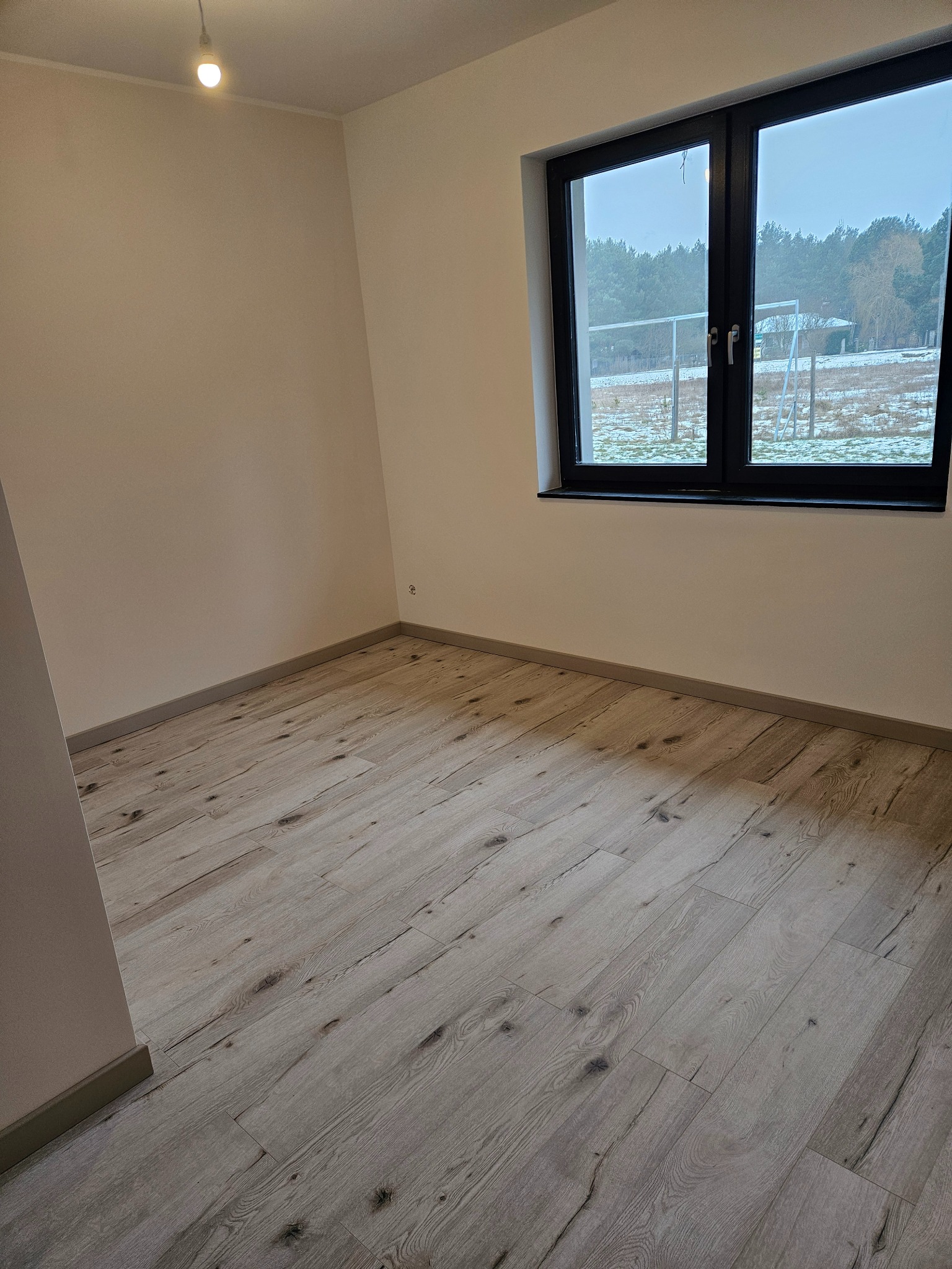 Jasne panele podłogowe w pustym pokoju z czarną ramą okienną i widokiem na zimowy krajobraz. Minimalistyczne wnętrze po remoncie.