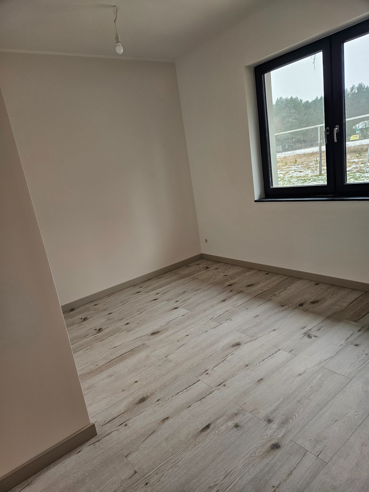 Jasne panele podłogowe w nowym pokoju z oknem na las. Ściany pomalowane na kremowo, surowa żarówka zwisa z sufitu. Minimalistyczne wykończenie wnętrza.