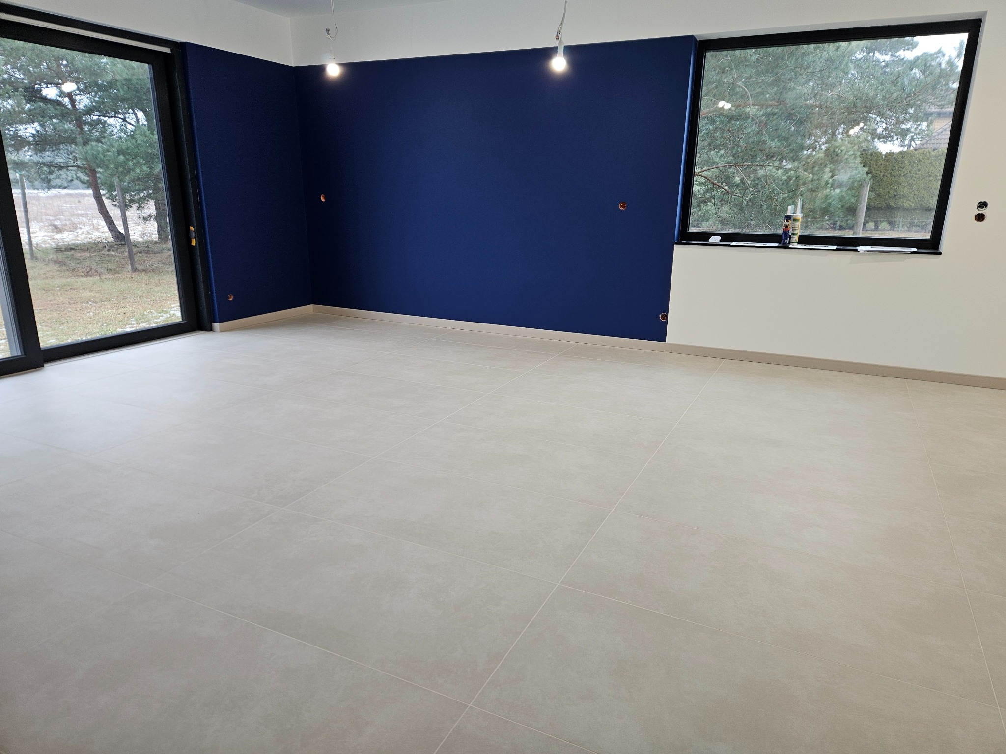 Wnętrze po remoncie: świeżo pomalowana ściana w odcieniach granatu, jasne płytki podłogowe, duże okno z widokiem na zewnątrz. Widoczne punkty elektryczne na ścianie.