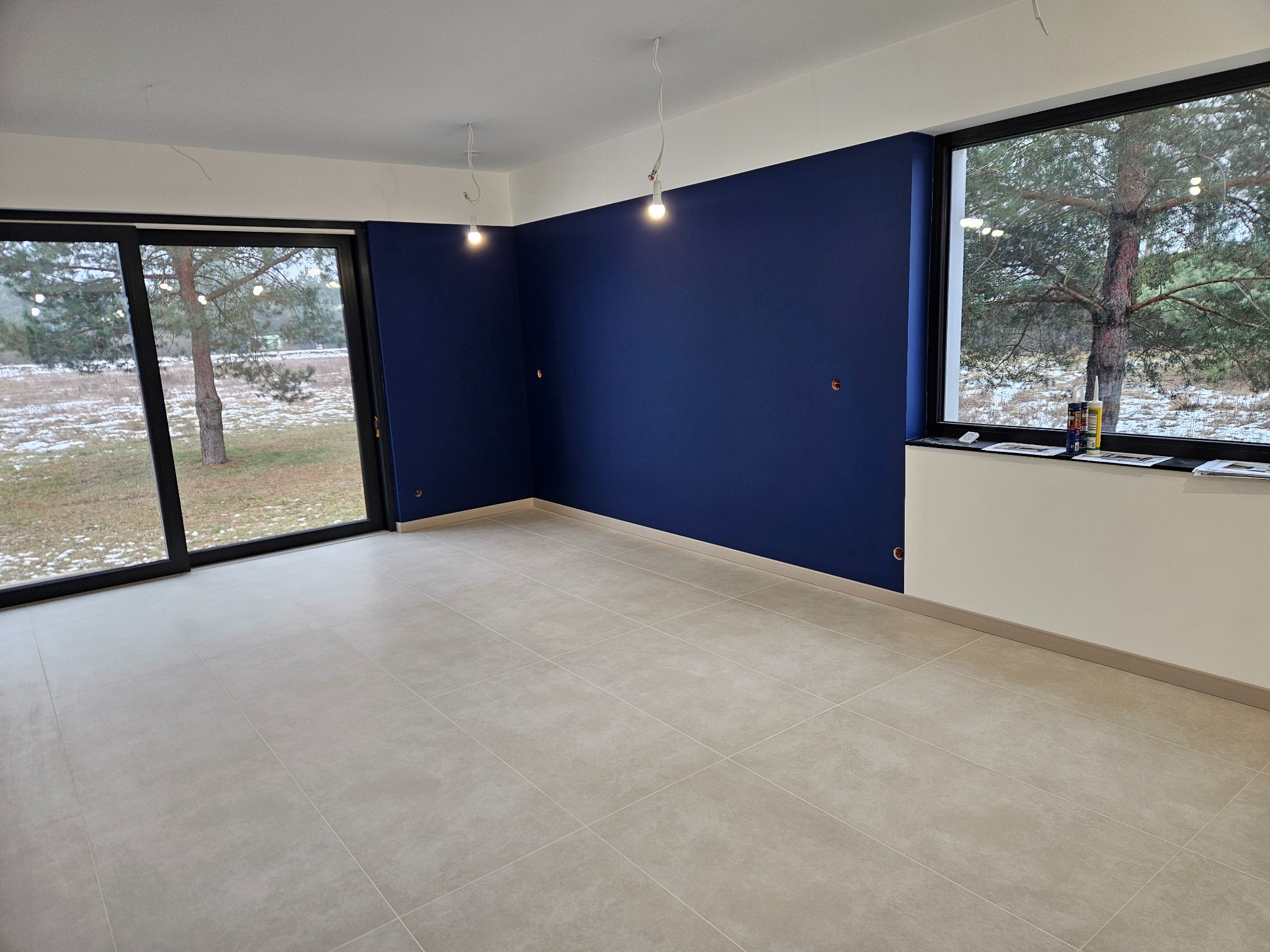 Wnętrze po malowaniu ścian na granatowo, z jasnymi płytkami podłogowymi i dużymi oknami z widokiem na zimowy krajobraz. Widoczne niedokończone oświetlenie sufitowe.