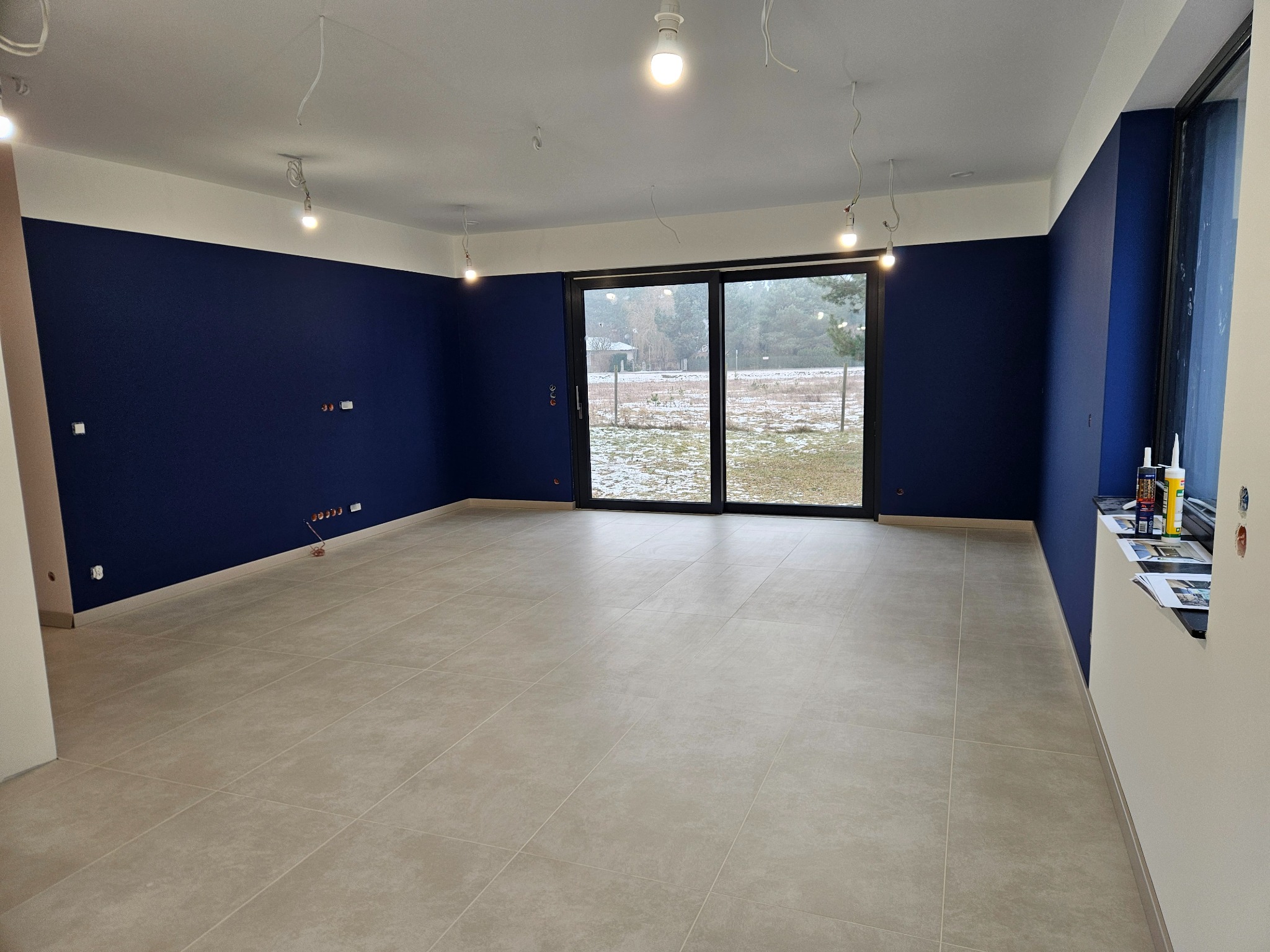 Wykończone wnętrze: granatowe ściany z białym pasem pod sufitem, beżowa podłoga z płytek, duże okno przesuwne. Widoczne niedokończone instalacje elektryczne.