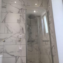 Marek Kowalski - Wykończona łazienka z kabiną prysznicową typu walk-in, wyłożona białymi płytkami imitującymi marmur, z widocznymi elementami instalacji elektrycznej i chromowaną armaturą.