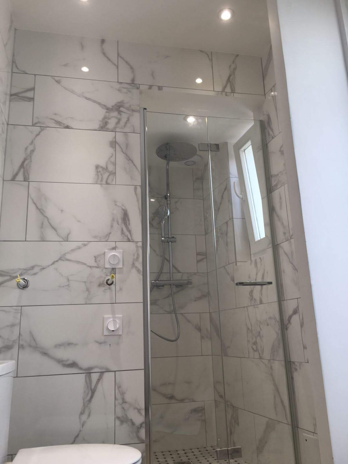 Wykończona łazienka z kabiną prysznicową typu walk-in, wyłożona białymi płytkami imitującymi marmur, z widocznymi elementami instalacji elektrycznej i chromowaną armaturą.