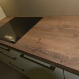 Fragment kuchni z blatem drewnopodobnym o wyraźnym usłojeniu i wbudowaną czarną płytą indukcyjną, widoczne szare fronty szafek z poziomymi uchwytami.