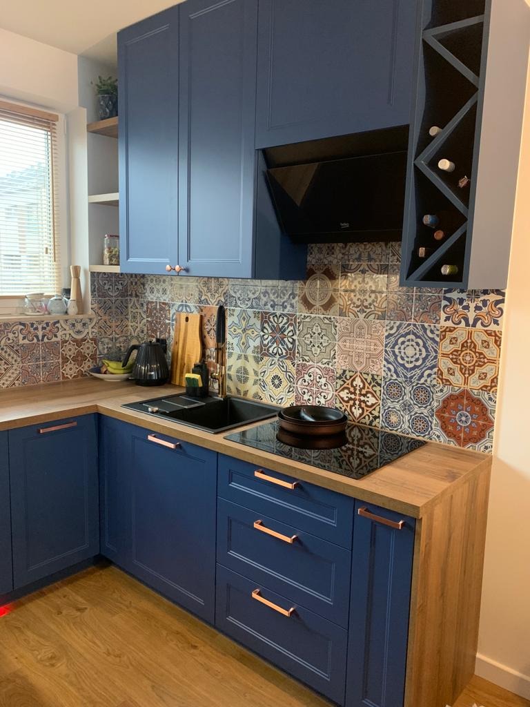 Niebieska zabudowa kuchenna z drewnianymi blatami i mozaiką na ścianie, widok na szafki górne z półką na wino i okap, nowoczesne uchwyty w kolorze miedzi.