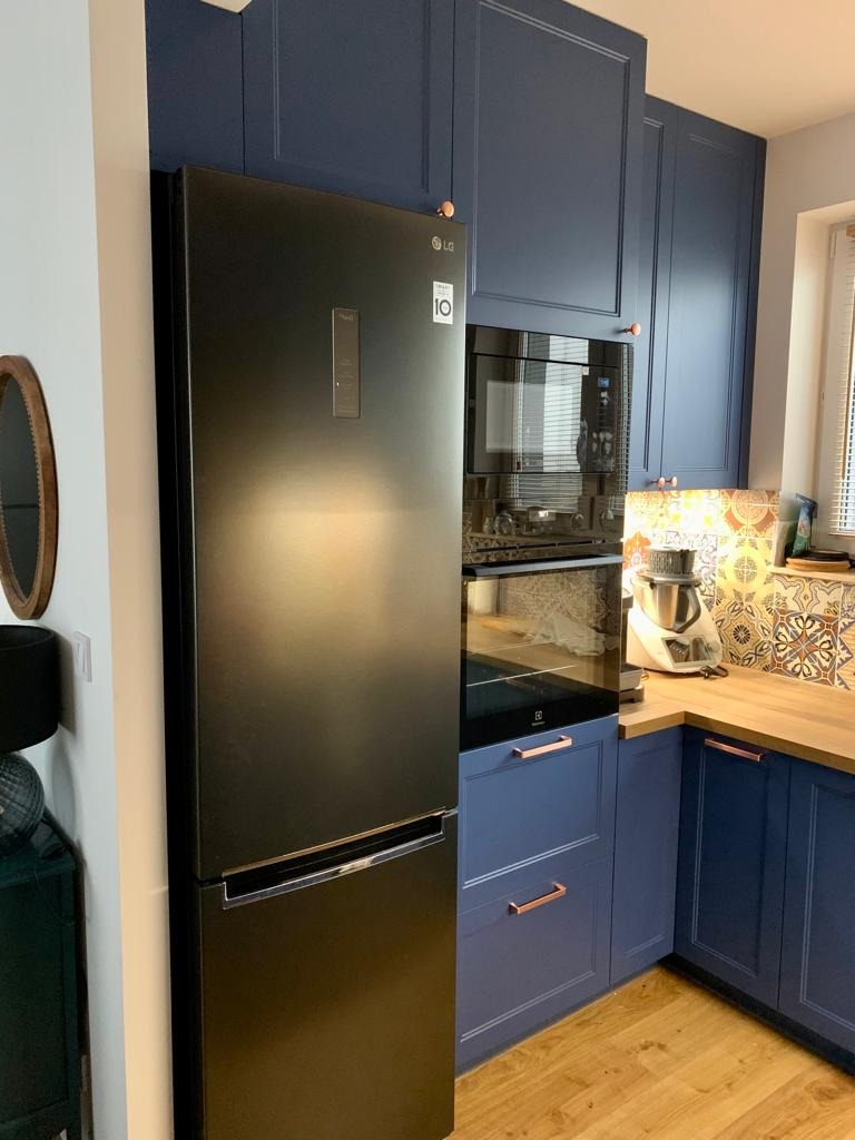 Granatowa zabudowa kuchenna z czarną lodówką, drewnianym blatem i miedzianymi uchwytami, widoczny fragment dekoracyjnej mozaiki nad blatem.