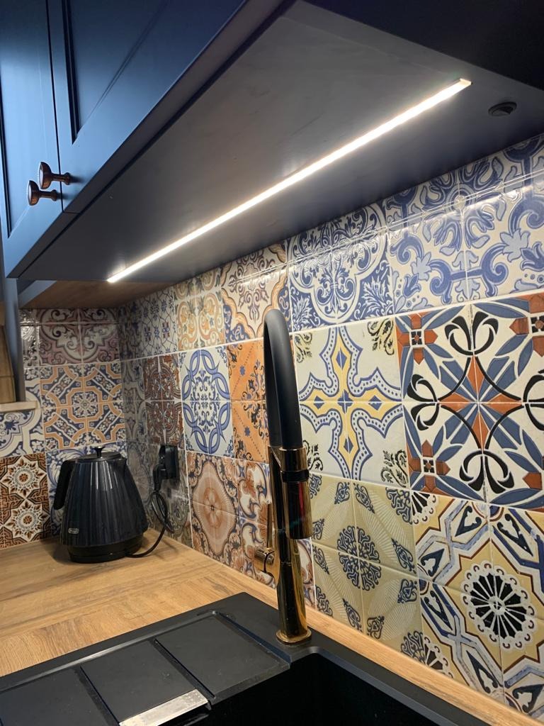 Fragment kuchni: ciemne szafki górne z oświetleniem LED podszafkowym, kolorowa mozaika na ścianie nad blatem, czarny czajnik elektryczny oraz bateria w kolorze złotym przy zlewozmywaku.