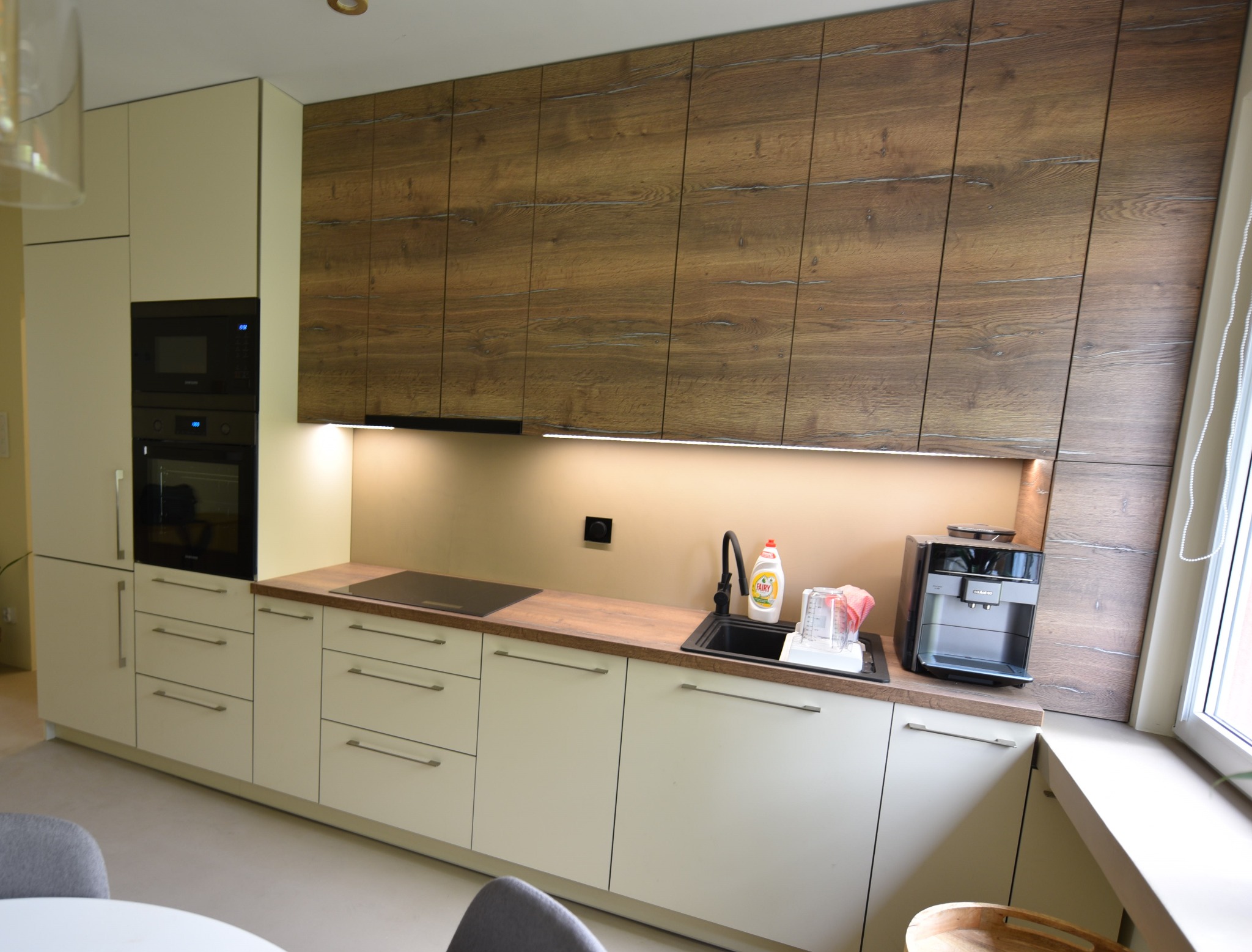 Nowoczesna kuchnia z jasnymi frontami dolnych szafek i górnymi szafkami w okleinie drewnopodobnej, czarnymi sprzętami AGD i blatem w kolorze drewna.