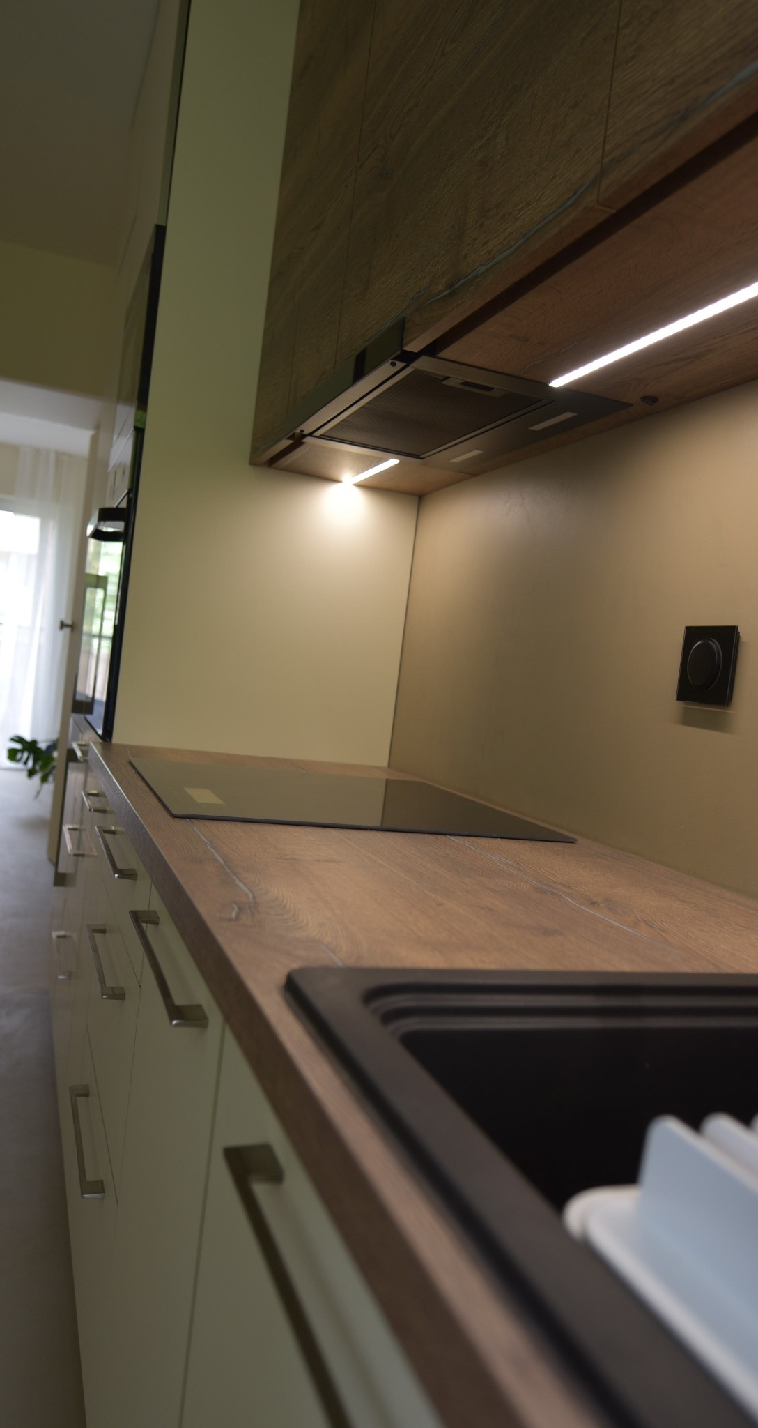 Fragment zabudowy kuchennej z drewnianym blatem, czarnym zlewem i płytą indukcyjną, oświetleniem LED pod szafkami i białymi frontami szafek z metalowymi uchwytami.