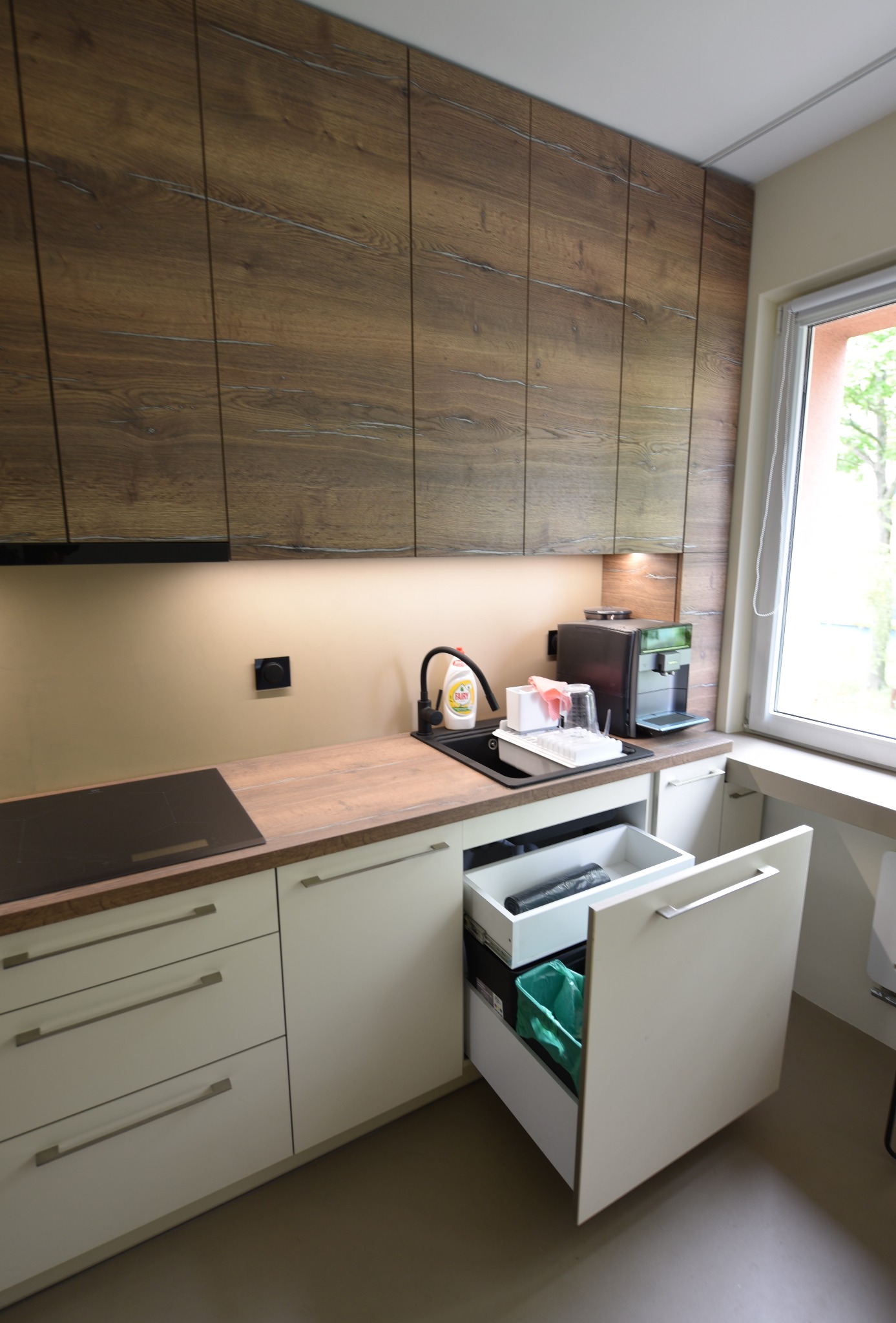 Nowoczesna kuchnia z drewnianymi szafkami górnymi i białymi dolnymi, otwartą szufladą na śmieci z workiem, czarnym zlewem i ekspresem do kawy przy oknie.