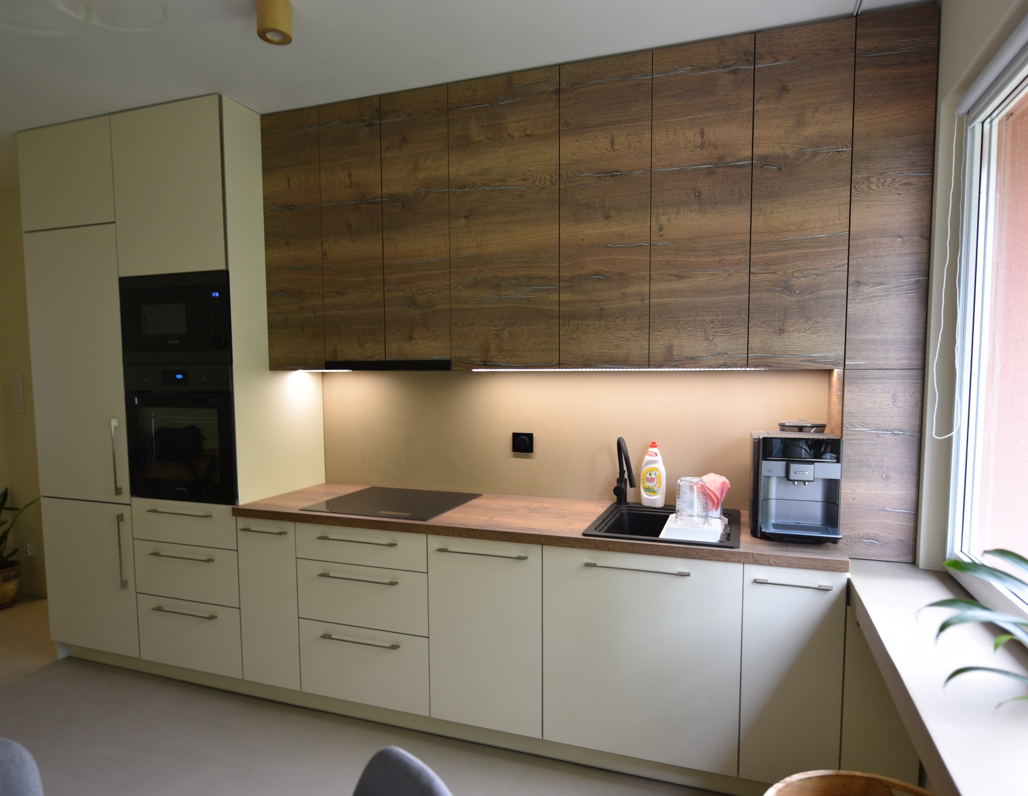 Nowoczesna kuchnia z białymi szafkami dolnymi i górnymi w kolorze drewna, z czarnym zlewem, płytą indukcyjną i ekspresem do kawy.