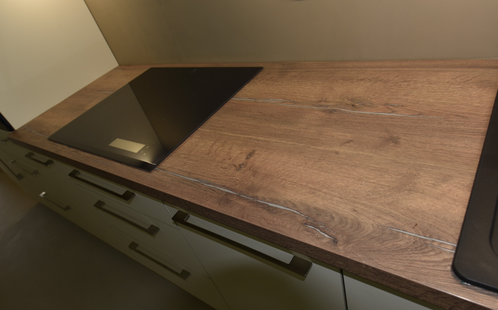 Fragment kuchni z blatem drewnopodobnym o wyraźnym usłojeniu i wbudowaną czarną płytą indukcyjną, widoczne szare fronty szafek z poziomymi uchwytami.