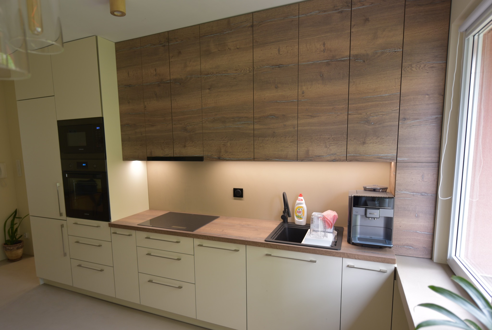 Nowoczesna kuchnia na wymiar z drewnianymi szafkami wiszącymi i jasnymi frontami dolnymi, wyposażona w czarny zlew, płytę indukcyjną i ekspres do kawy.