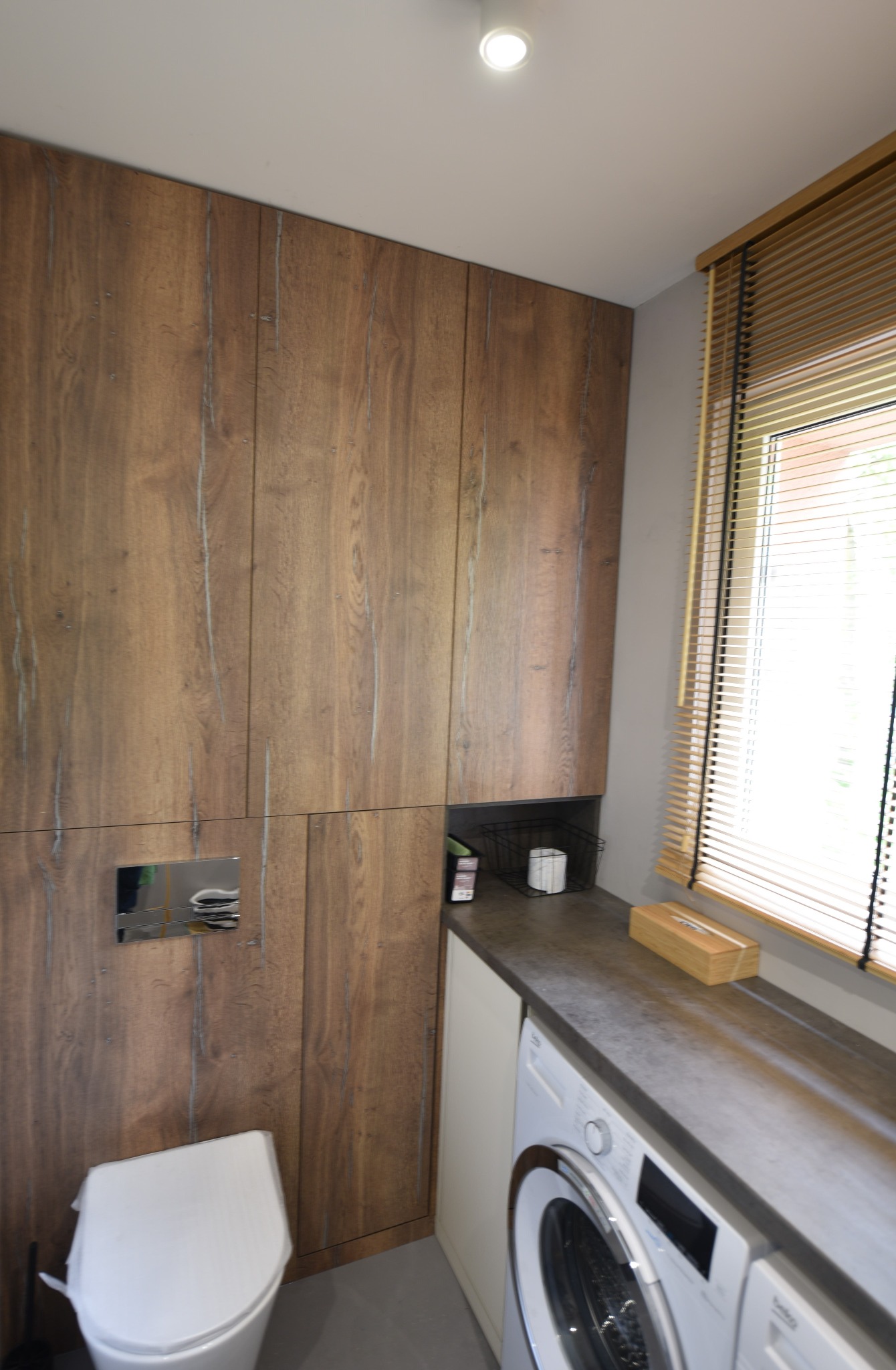 Drewniane szafki łazienkowe z ukrytą spłuczką, blat z szarego konglomeratu nad pralką, drewniane żaluzje w oknie.