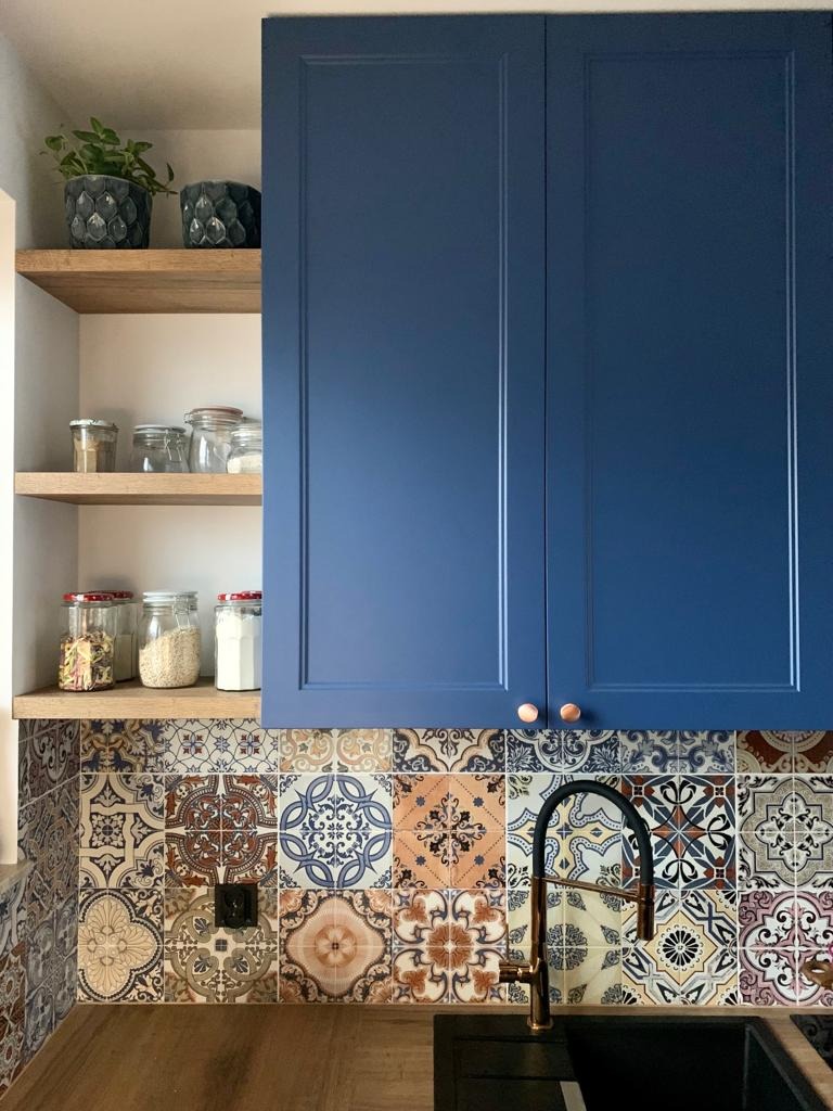 Niebieskie szafki kuchenne nad blatem z drewnianym wykończeniem i mozaiką wzorzystych płytek, czarna bateria umywalkowa, otwarte półki z zapasami i roślinami.
