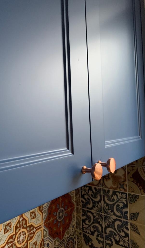 Fragment błękitnej szafki kuchennej z miedzianymi uchwytami na tle wzorzystych płytek.