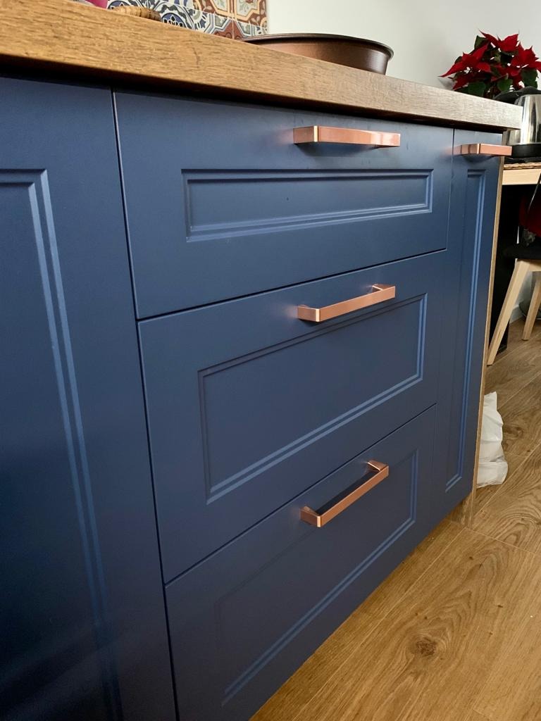 Granatowe szafki kuchenne z frezowanymi frontami i miedzianymi uchwytami, widok z bliska na szuflady i fragment drewnianego blatu.
