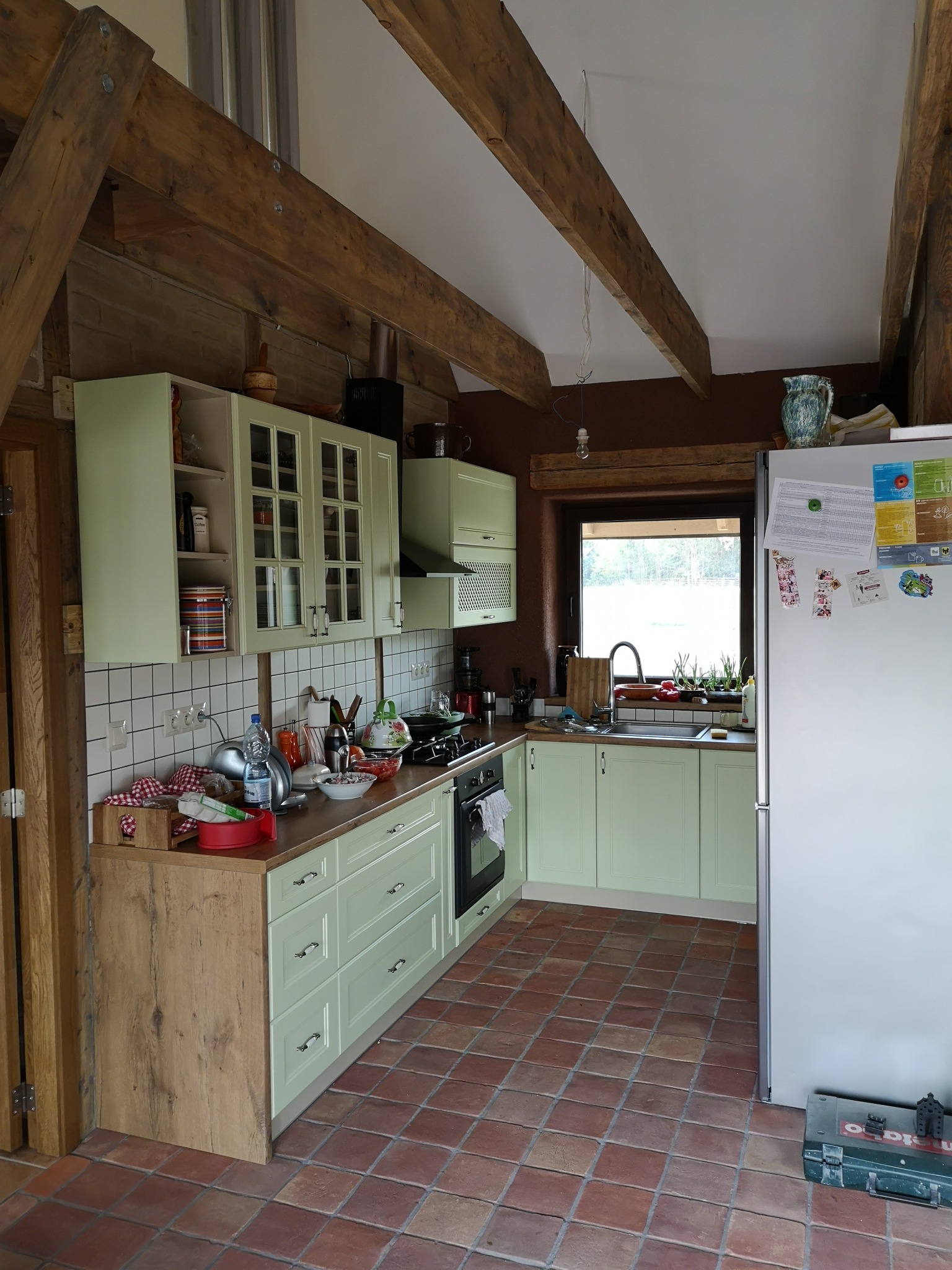 Kuchnia w stylu rustykalnym z drewnianymi belkami stropowymi, jasnymi szafkami i terakotową podłogą; na blacie widoczne naczynia i akcesoria kuchenne.