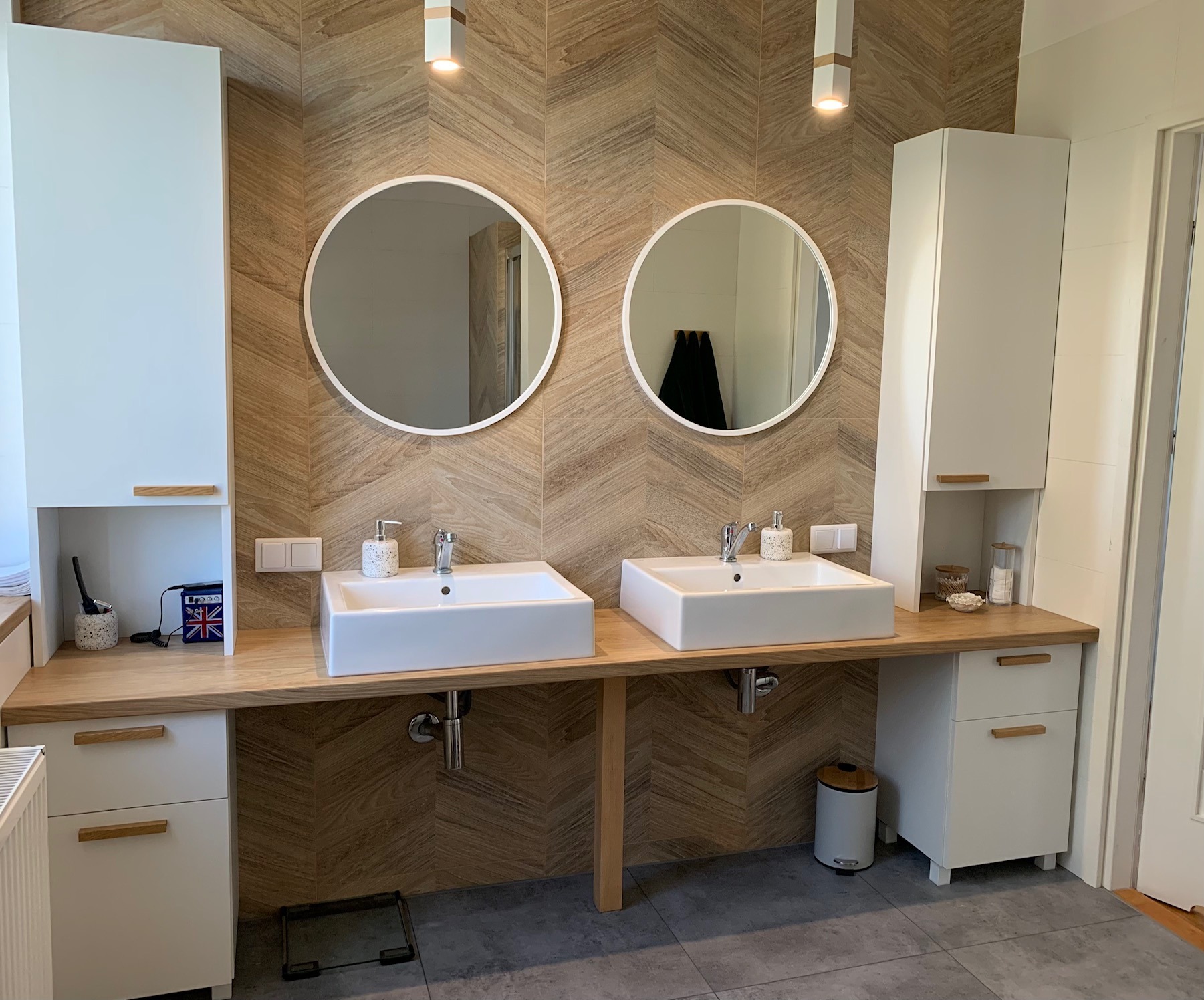 Nowoczesna łazienka z dwoma umywalkami nablatowymi, okrągłymi lustrami, drewnianym blatem i szafkami w kolorze białym, podświetlona wiszącymi lampami.