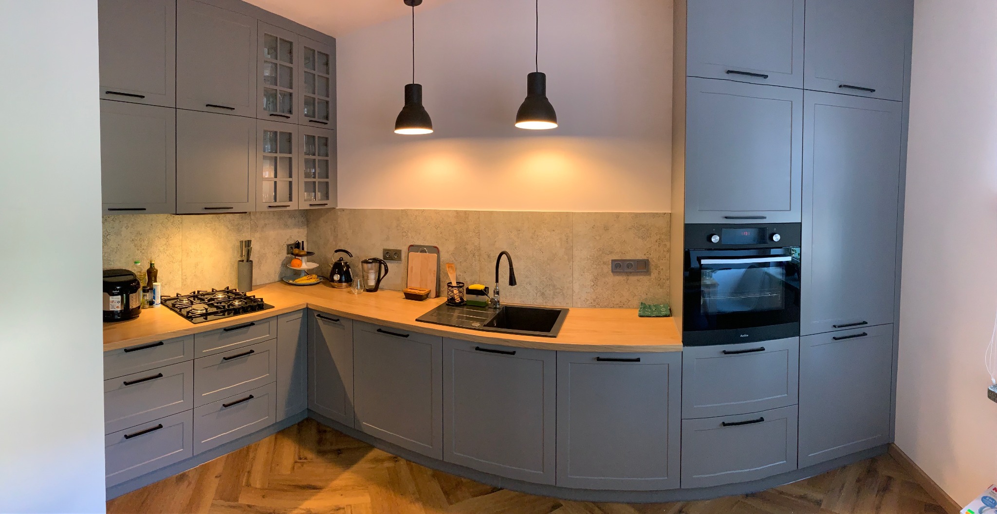 Szara kuchnia na wymiar z drewnianym blatem, czarnym zlewem i baterią, oświetlona dwoma wiszącymi lampami w stylu industrialnym, z parkietem ułożonym w jodełkę na podłodze.